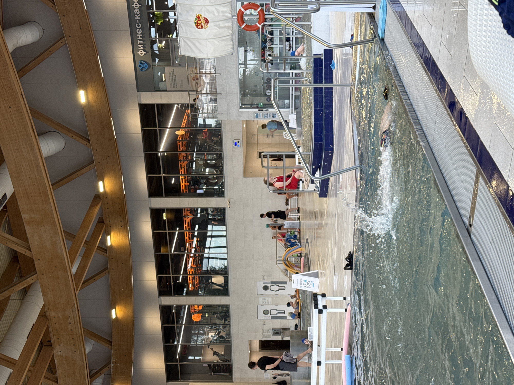 This is an indoor swimming pool facility, likely part of a fitness center, located in Sovkhoz imeni Lenina, Russia. The architecture features a high, curved wooden ceiling with exposed beams and ventilation systems, illuminated by modern light fixtures. Large windows line the upper level, offering views into a gym area with exercise equipment.
In the foreground, a swimming pool with clear, rippling water occupies a significant portion of the frame. Splashes are visible, indicating activity. A person is partially submerged in the water, appearing to be swimming or engaging in aquatic exercise. Along the edge of the pool, there are metal railings and tiled surfaces.
In the middle ground, there is a shallower area of the pool with tiled steps and seating. Several people are present here, some sitting or standing, and a few appear to be children in the water, possibly participating in a lesson or supervised play, surrounded by inflatable pool toys. A man in a black t-shirt and shorts stands on the tiled area, holding a towel. Signs are visible, including one with a depiction of a person falling, suggesting safety instructions.
The background reveals the gym area visible through the windows, with rows of treadmills and other exercise machines. A café or reception area is also indicated by signage. A life preserver is hung on a wall near a section of the pool. The overall environment appears clean, modern, and functional, designed for both recreation and fitness.