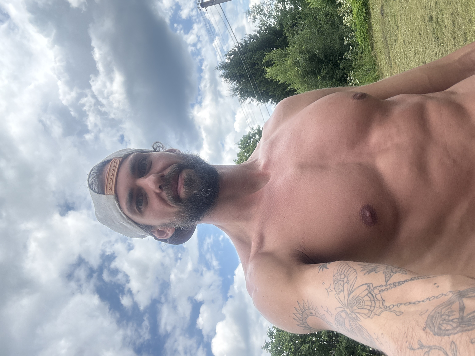 This media file is a selfie taken outdoors, likely during the daytime. The subject is a man with a beard and a baseball cap, who is shirtless. His torso and arm are visible, and he has tattoos on his arm, including a butterfly. The background consists of a cloudy sky, trees, and a grassy area, suggesting a rural or suburban setting. There are also power lines visible in the background. The man's expression is neutral. No text is visible in the image. The location context is Kuti, Russia.