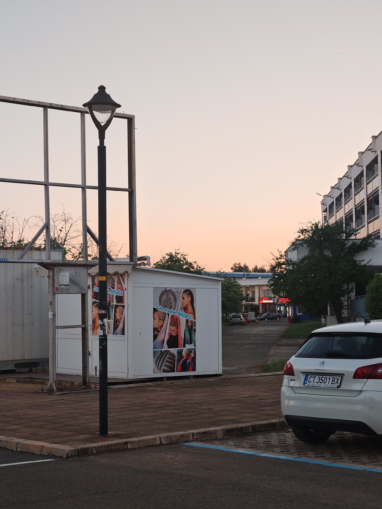 Here's a forensic description of the image:
The image depicts an outdoor scene in what appears to be a parking area near a building complex. The sky is tinted with the colors of a sunrise or sunset. The foreground is dominated by a paved area, with parking space markings visible.
A white Peugeot 308 is parked, with its rear facing the camera. The license plate number is visible: CT.3501 BX.
To the left, there is a small, white portable structure covered with posters advertising a hair braiding service. A lamp post stands in front of the structure. Behind the portable structure is a large, empty billboard frame.
In the background, a multi-story building with a row of balconies is visible. Further in the distance, there is a restaurant with a sign that includes the word "SUPER." Several cars are parked in this area.
Based on the information present in the image, specifically the car's license plate, the location is likely Nesebar, Bulgaria.