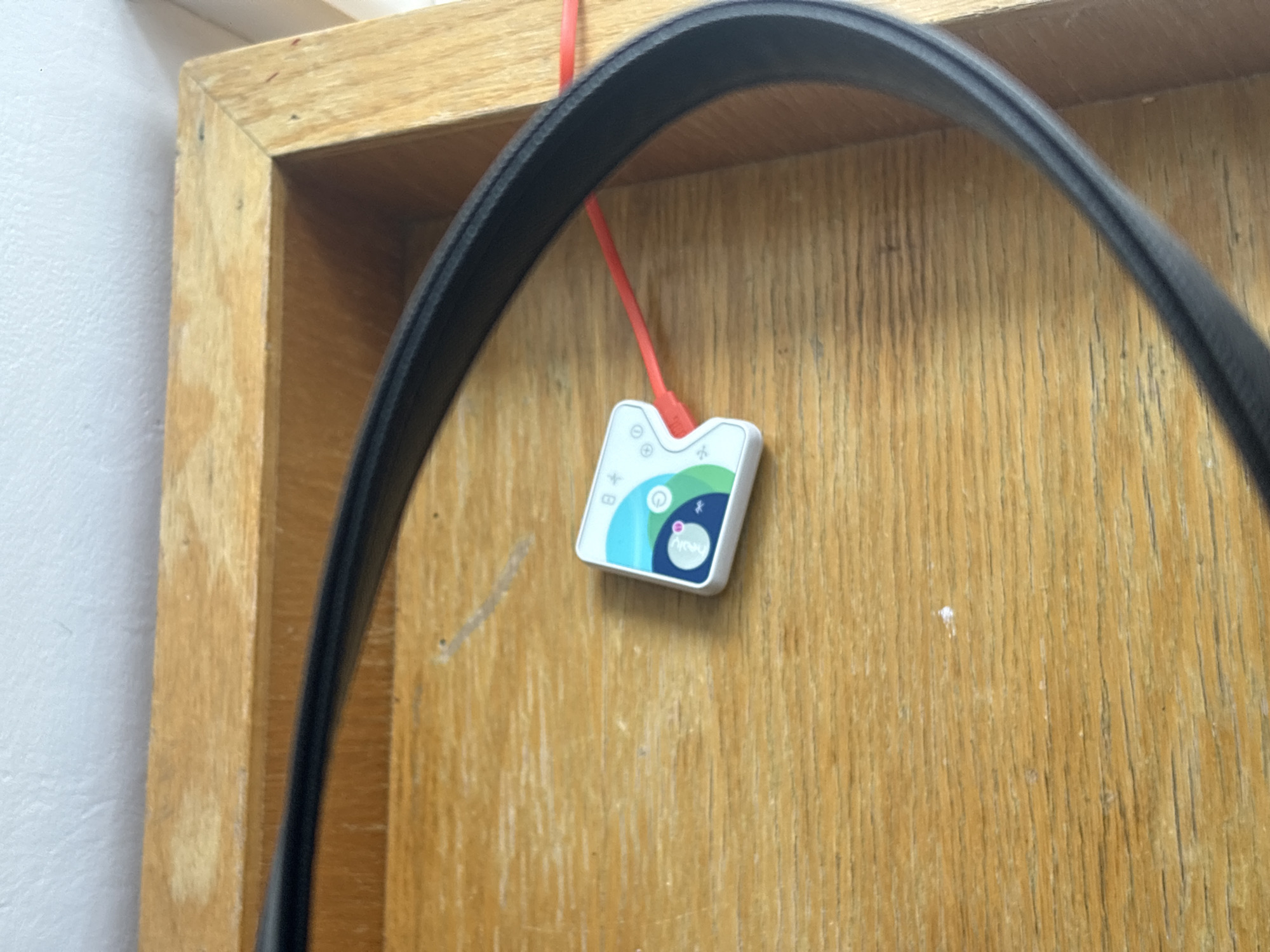 The image depicts a wooden cabinet or shelf with a white square device attached to it by a thin red cord. The device has a colorful design with green, blue, and white elements and features the word "Nevy" on it. A black strap or belt is partially visible in the foreground, obscuring some of the scene. The wooden cabinet has a light brown color with vertical grain patterns. The wall behind the cabinet is painted white. The location is likely indoors. There is no information available to confirm the location as Mexico City, Mexico.