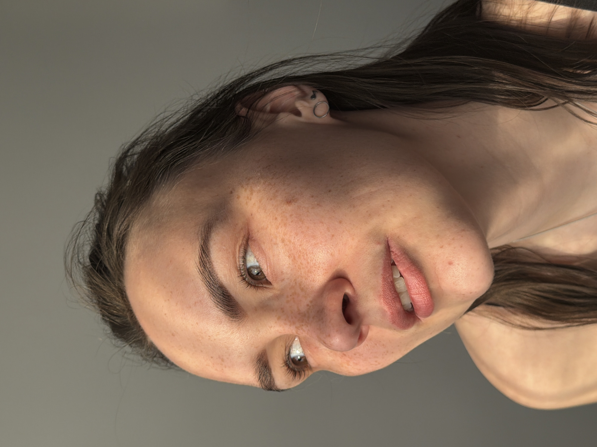 A close-up portrait of a woman with brown hair and freckles is presented against a gray background. The woman is partially turned, and her face is in focus. The image shows her eyes, nose, mouth, and the side of her face. A small earring is visible in her ear. The lighting suggests a natural source, highlighting the details of her skin and hair. No specific information indicates the location is Chaikovsky, Russia.