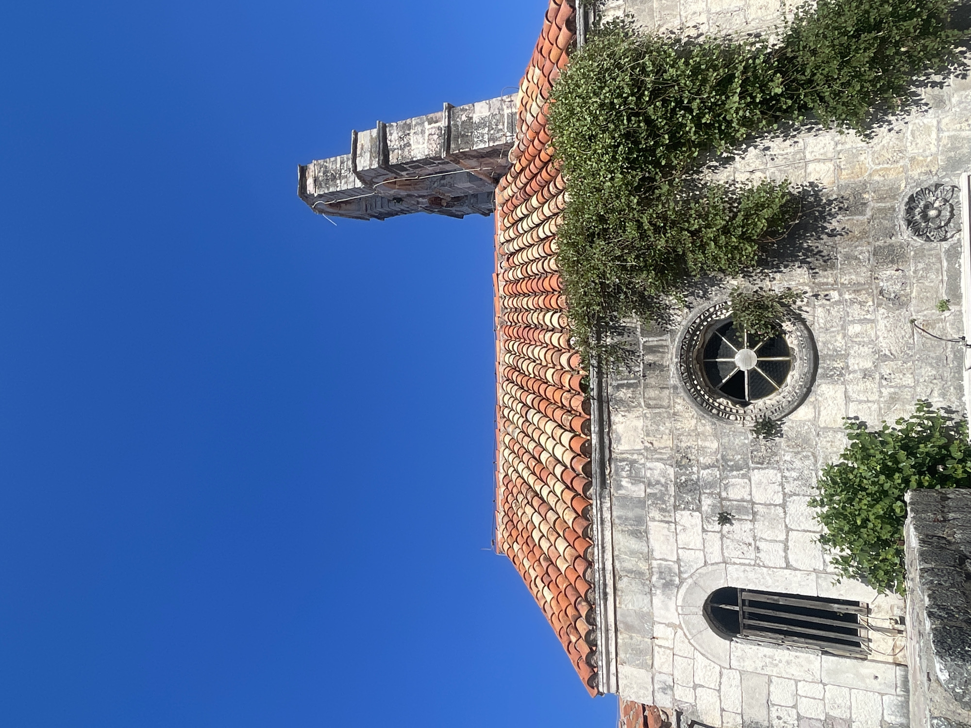 The image shows an exterior view of a building against a clear blue sky. The building features a terracotta tile roof, a round window with a decorative surround, a lower window with a boarded-up appearance, and stone construction with vegetation growing on the walls. The architectural style suggests a historical or possibly medieval building, likely a church or other significant structure. The location is Unknown, Unknown.