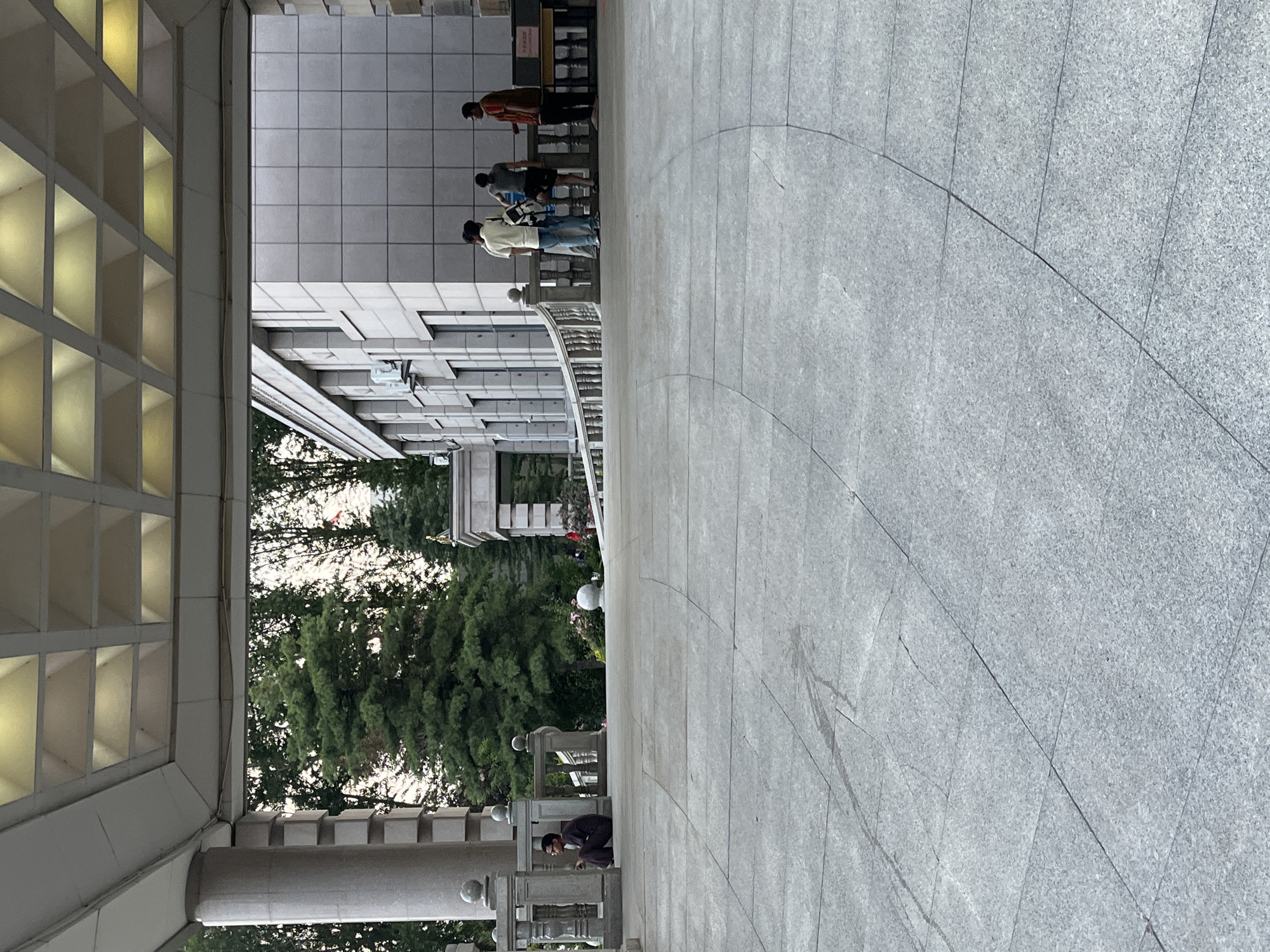 A paved outdoor area is shown under a building's overhang. The overhang has a textured, grid-like ceiling. To the left, a person in dark clothing sits on the ground near a stone column and railing. In the middle ground, several people stand near a white railing. Behind them is a modern building with windows and air conditioning units. Lush green trees are visible in the background. The paving stones are large and grey, with some dark marks and cracks. The overall scene suggests an urban environment, potentially a public space or the entrance to a building.
