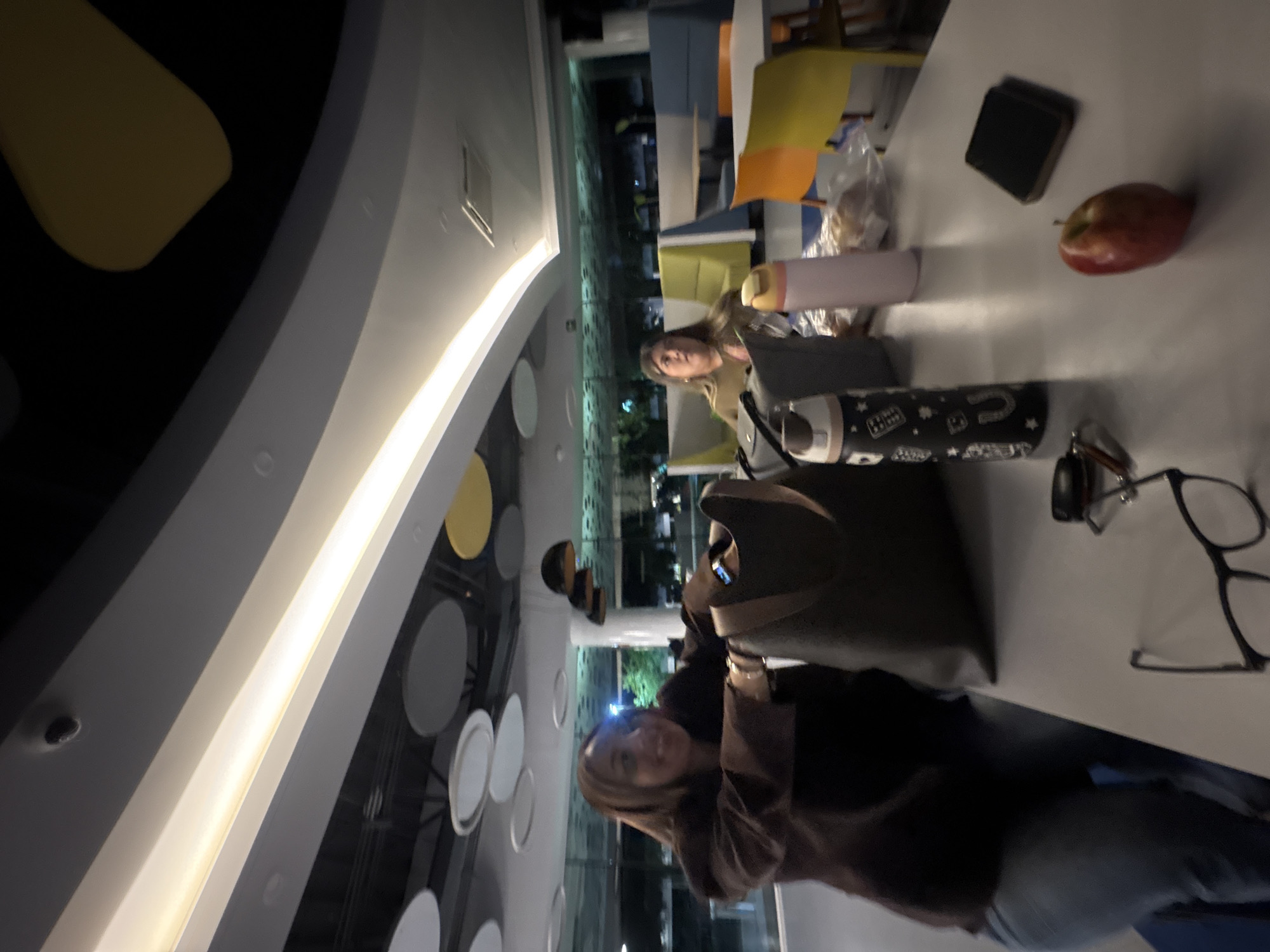 The image captures an indoor scene at night, likely in a modern café, lounge, or common area in Puebla, Mexico. Two women are seated at a table, facing the camera.
The woman closer to the viewer, positioned on the right side of the frame, has light brown hair and is wearing a dark, possibly textured, top and blue jeans. She has a faint smile and is looking directly at the camera. Her upper torso is partially obscured by a dark bag placed on the table in front of her. The second woman, further back towards the center-left, also has light brown hair and is wearing a lighter, possibly khaki-colored, top. She is looking towards the camera with a neutral expression.
The table between them holds several items: a dark brown or black handbag, a light pink water bottle, and a black water bottle adorned with white designs including stars, a horseshoe, and the word "LUCKY." Other objects on the table include a pair of black-framed glasses, a red apple, a stack of black coasters, and some crumpled clear plastic, possibly from a snack.
The background shows large windows looking out onto a dark exterior with distant lights, confirming the night-time setting. The interior features contemporary lighting, including a bright strip light along a curved architectural feature on the ceiling/wall, and circular decorative elements. The overall atmosphere appears relaxed and social, with the women casually seated and aware of the camera.