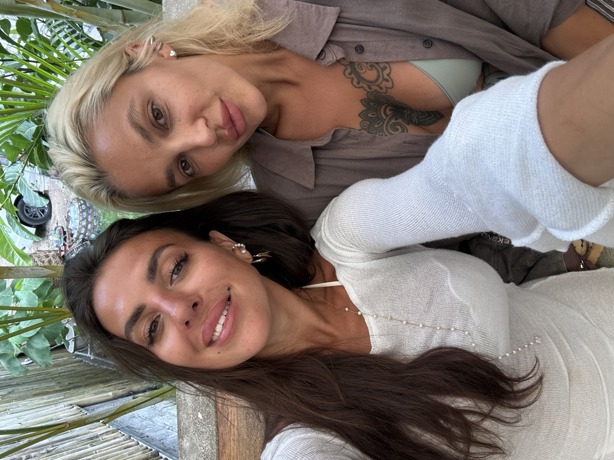 Two women are taking a selfie outdoors in Kuta Selatan, Indonesia. The woman on the left has long, dark brown hair and is wearing a white, long-sleeved top with a pearl necklace. The woman on the right has blonde hair and is wearing a grey open shirt over a light green top. She has a tattoo on her chest. They are seated, with lush green foliage and bamboo visible in the background, suggesting a tropical setting.
