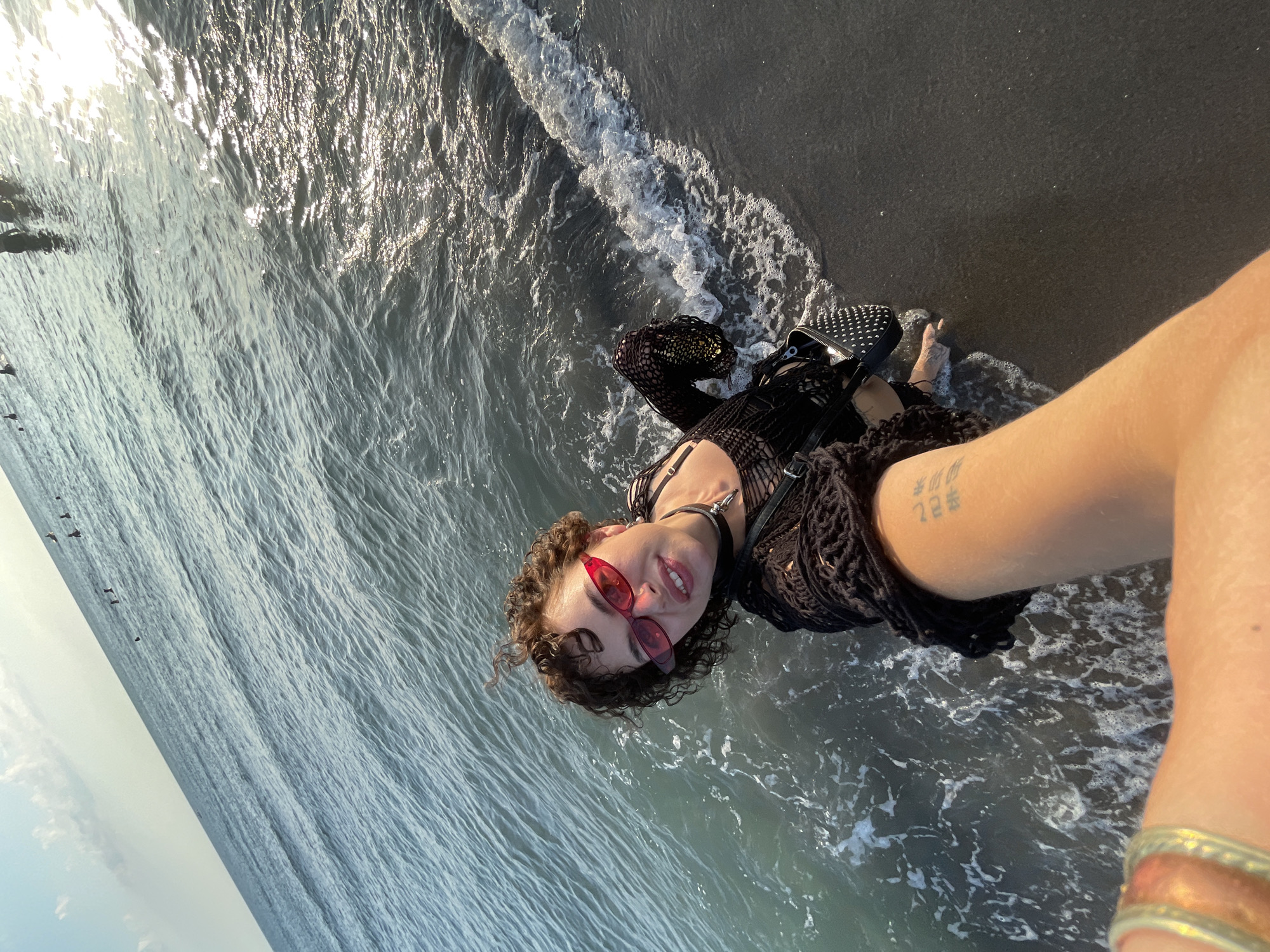A high-angle, wide shot shows a person taking a selfie at the edge of the Adriatic Sea in Donji Štoj, Montenegro. The person is wearing red-tinted sunglasses and a black mesh top. They are standing in shallow water, with waves lapping at their feet. The beach is dark sand, and the sea is a deep blue-green. In the distance, several other people can be seen in the water. The sun is setting, casting a warm glow over the scene.