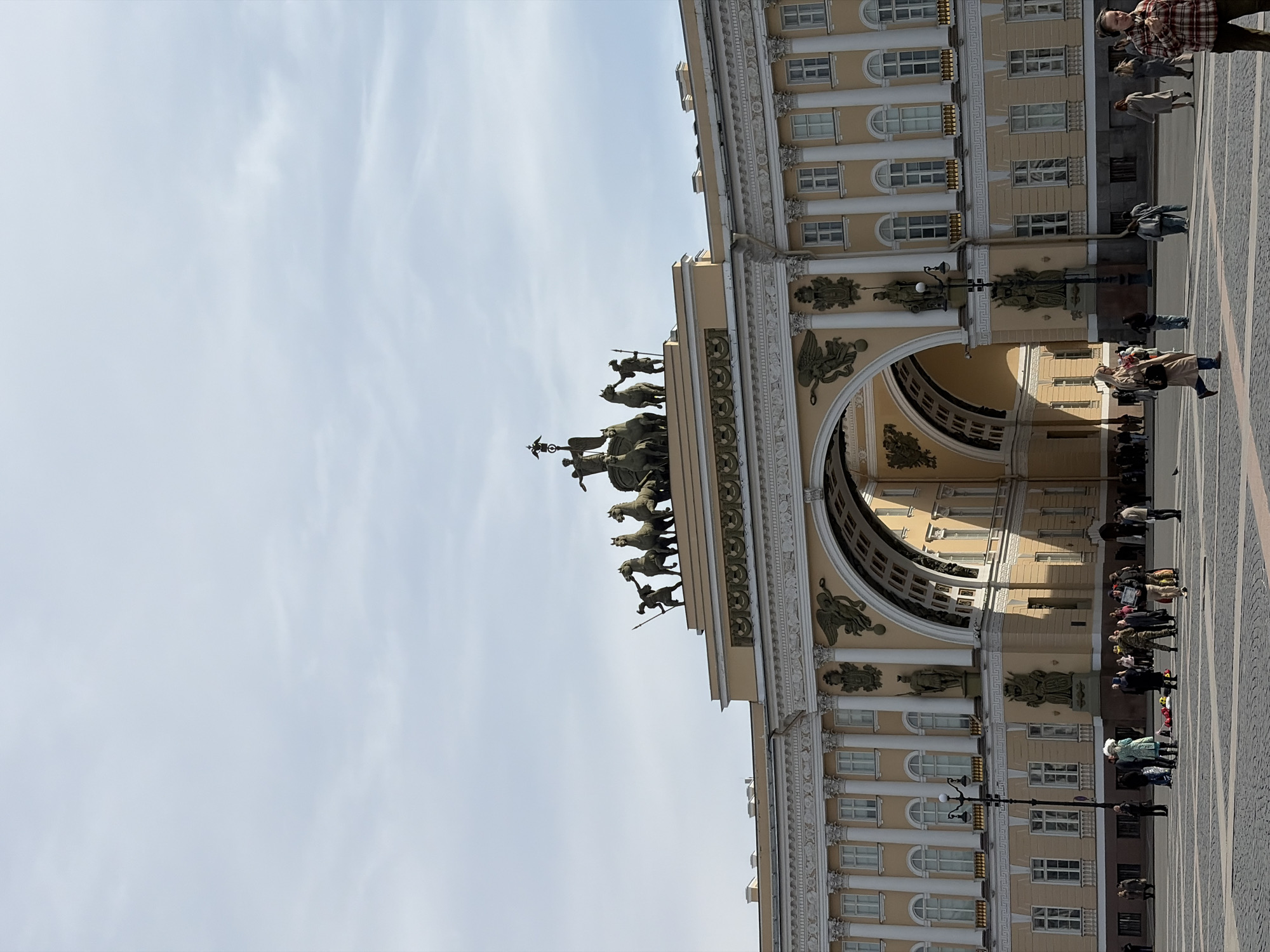 The image is a vertical shot of the General Staff Building in Saint Petersburg, Russia, taken at ground level. The camera is oriented to capture the building’s facade and the surrounding square.
The building’s facade is predominantly cream-colored, with classical architectural elements, including columns, arched windows, and decorative statues. The upper portion features a triumphal arch topped with a sculptural group depicting figures and horses.
Several pedestrians are visible in the square, walking and standing near the building. The square itself appears to be paved with cobblestones or similar materials. The sky is visible to the left of the building, appearing light blue with scattered clouds.
The light indicates it is daytime. The image lacks any signs of recent modifications.