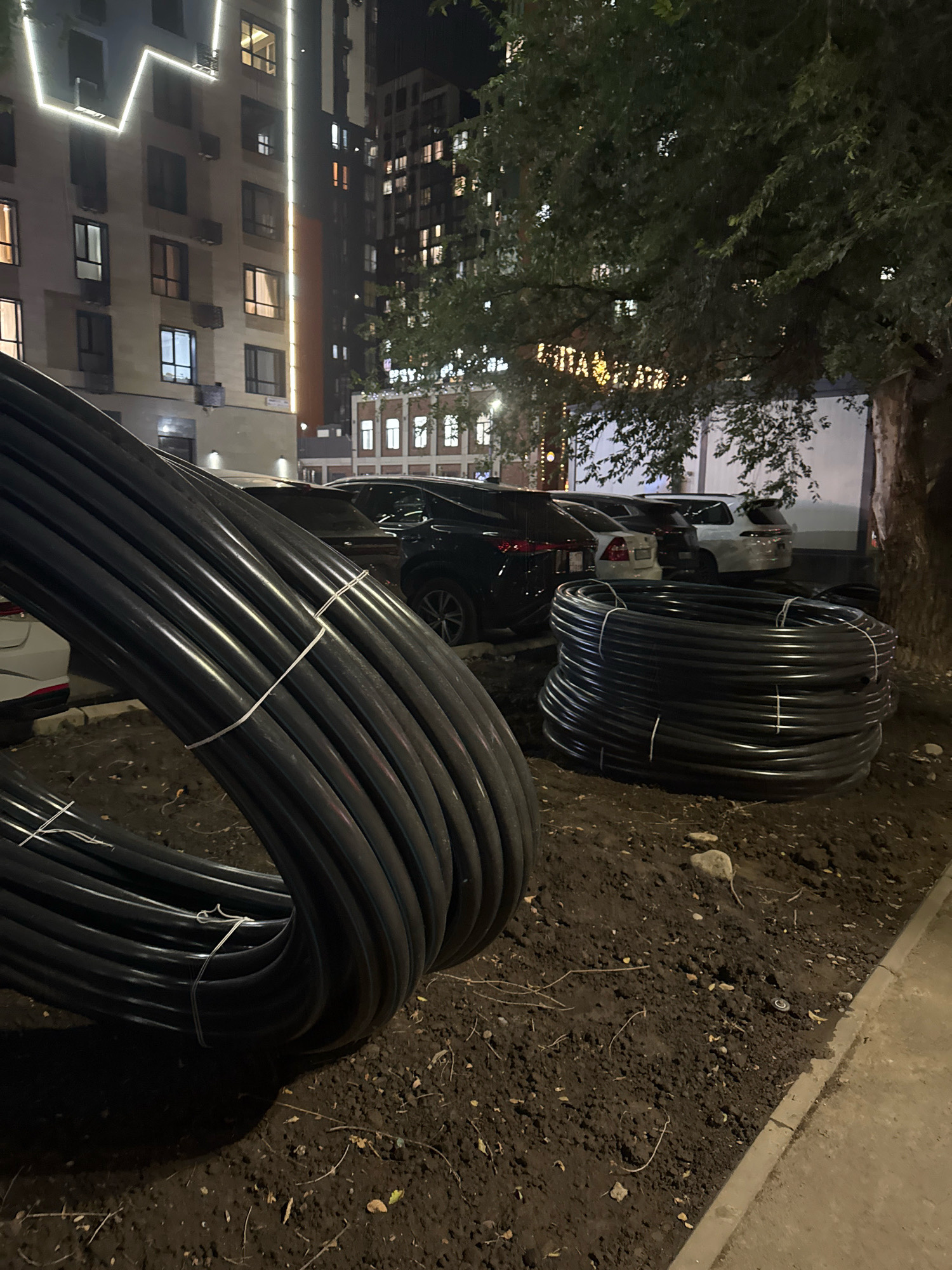 In Almaty, Kazakhstan, at night, a close-up shot shows large coils of black plastic piping laid on the ground in front of a multi-story residential building. The building has illuminated windows and decorative lights on its facade. Several cars are parked in the background, and a large tree with dark green foliage is to the right of the frame. The ground is covered in dark soil with scattered leaves and small stones. The piping is coiled and secured with white zip ties.