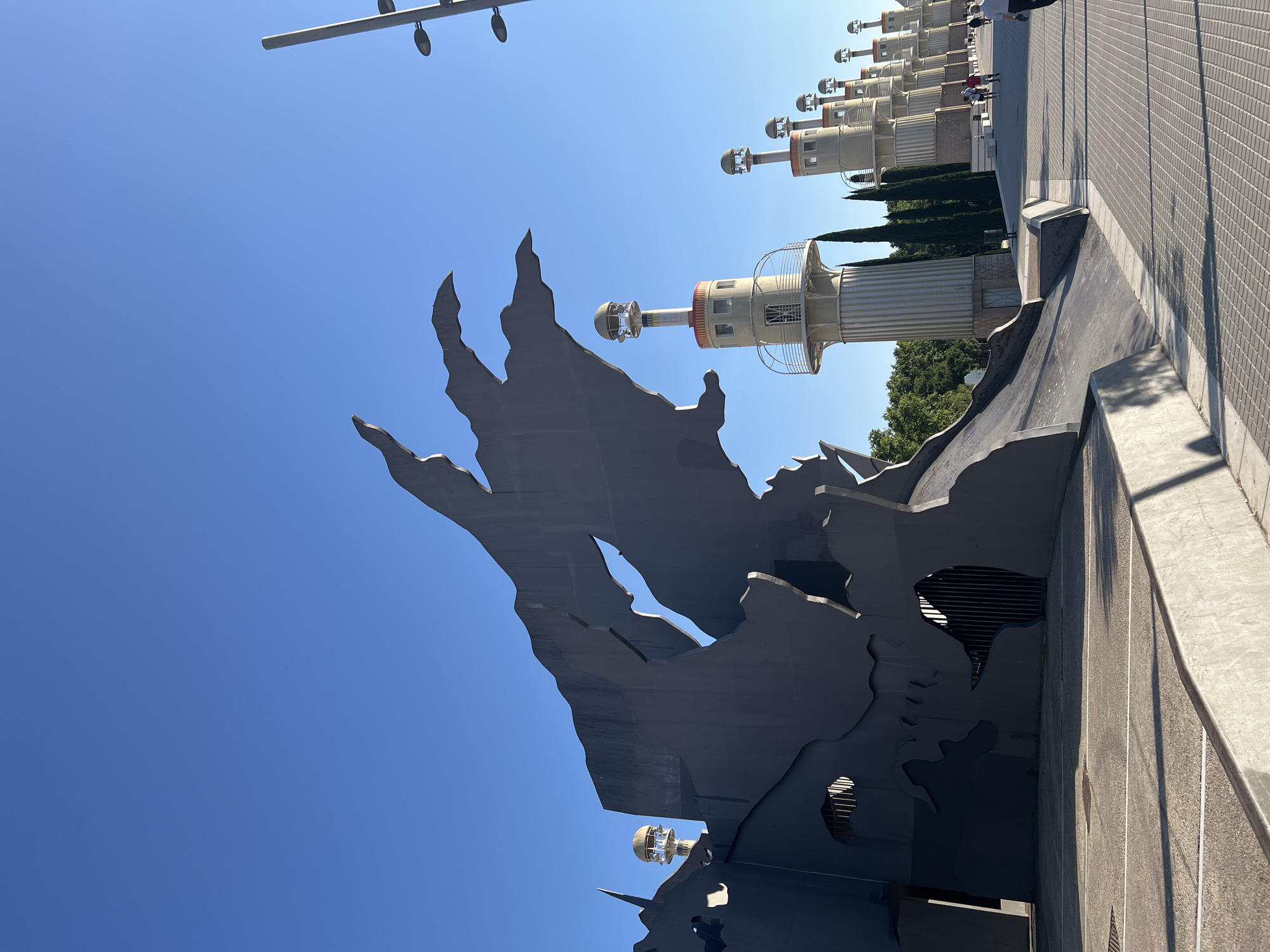 The image shows a street scene in Barcelona, Spain, on a sunny day. The camera is tilted upwards, capturing a large, dark, decorative structure in the foreground, that looks like a map. Behind the structure, a row of futuristic-looking towers extends into the distance. The towers are cylindrical with multi-tiered segments, a circular upper section, and topped with a round, glass-like dome. The sky is clear and blue. The towers are located near a pedestrian walkway. Some people can be seen in the background near the towers.