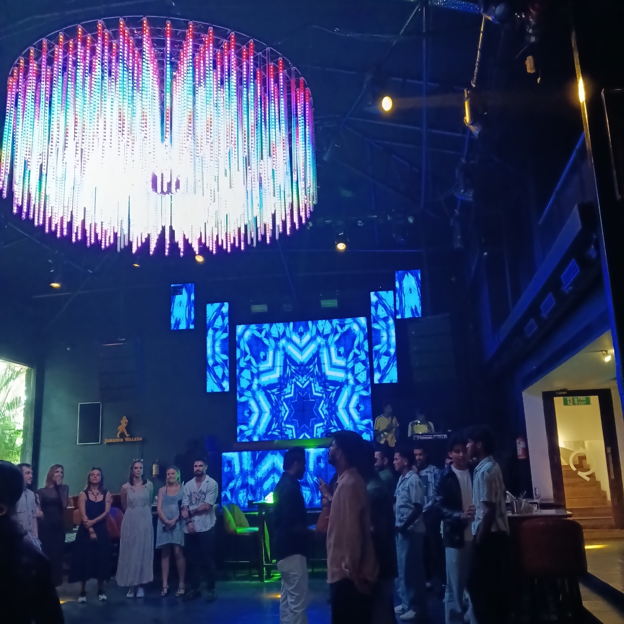 The image shows a nighttime scene inside a venue in Sinquerim, India. Overhead is a large, circular chandelier with rows of colorful LED lights hanging down. Behind a gathering of people are several large LED screens displaying blue geometric patterns. Some people are standing facing the screen, while others are engaged in conversation. Some are dressed in casual attire. Towards the back of the room, two people can be seen playing instruments. In the background, there is a bar area with a staircase leading upwards. A "Johnnie Walker" sign is visible. The lighting is predominantly blue, creating a nightclub-like atmosphere.