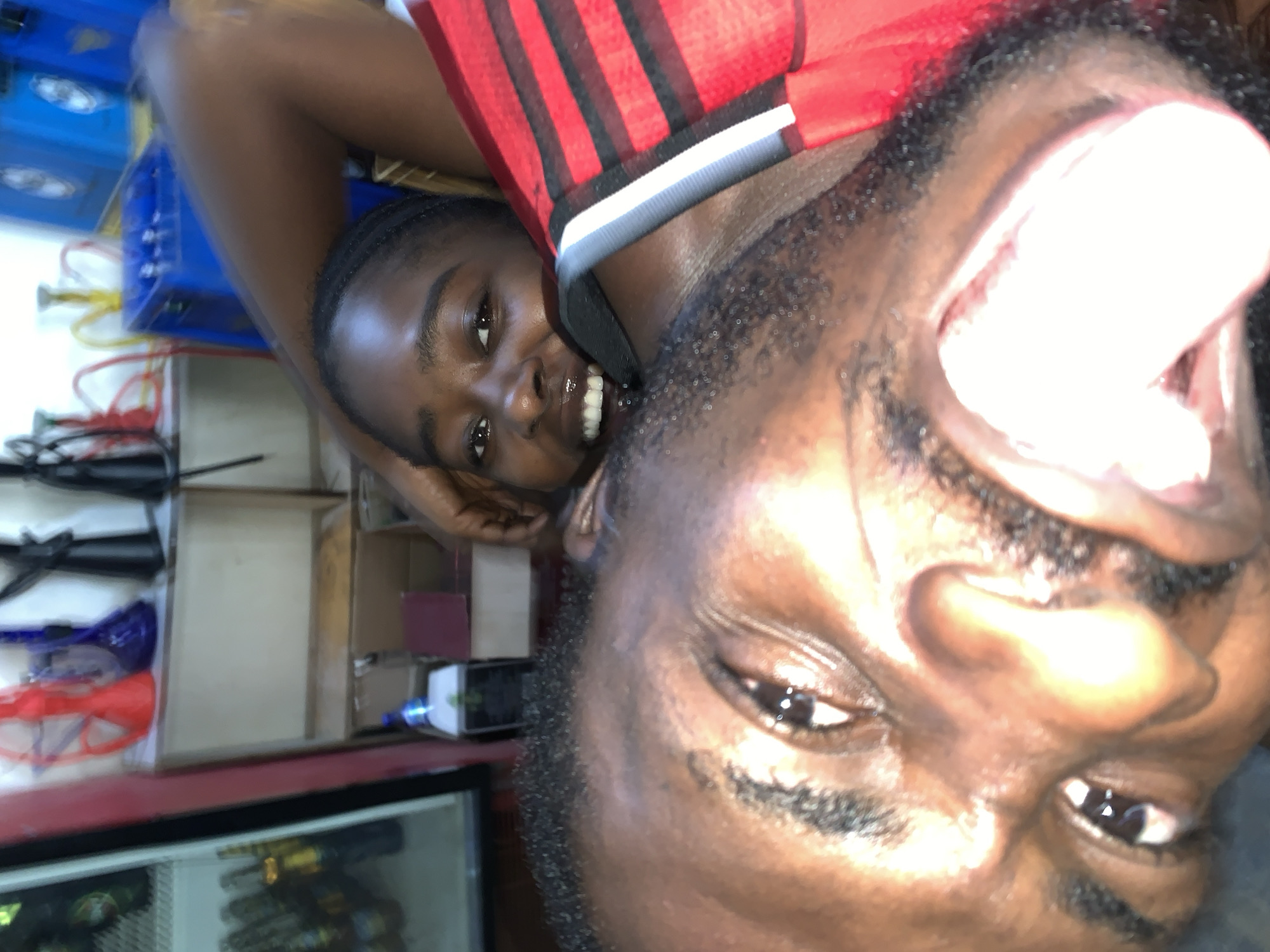 This image, rotated counter-clockwise, shows two people in what appears to be a small commercial establishment, possibly a convenience store. One person, with dark skin and short braided hair, is smiling and looking at the camera. The other person, with a beard, is positioned in the foreground. The people are wearing a black, red and white striped shirt.
In the background, there is a cooler with beverages, shelves stocked with items, and blue containers, all of which suggest a commercial setting. It's difficult to verify the location of New Stadium, Nigeria, without further details or external information.