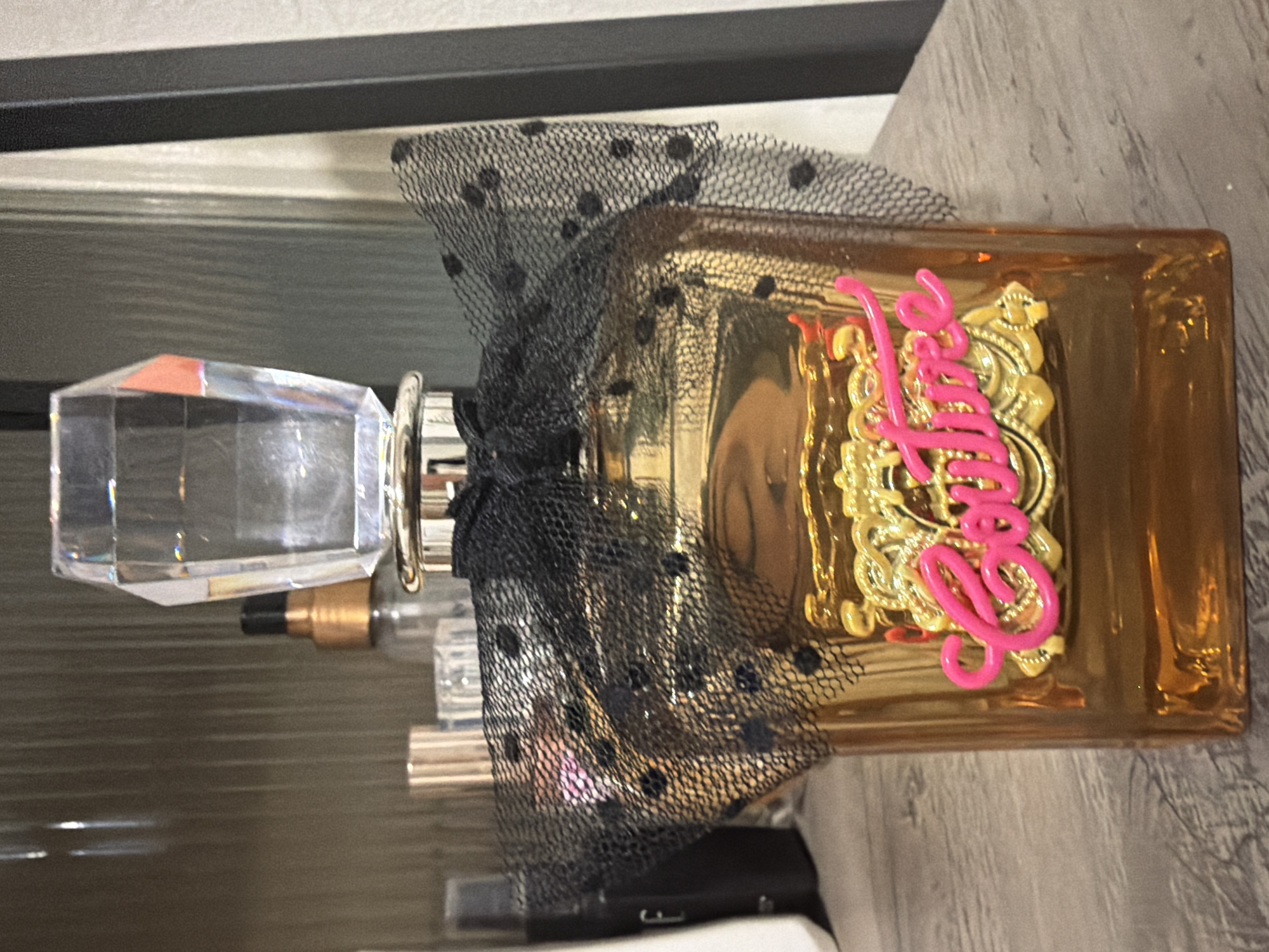 The image shows a bottle of Juicy Couture perfume on a gray surface, likely a dresser or vanity. The bottle is amber-colored glass with a clear, multifaceted crystal cap. A black polka-dot mesh ribbon is tied around the neck of the bottle. The perfume's name, "Juicy," is written in pink script on a gold emblem on the bottle. The environment is domestic, but the location within the city of London, United Kingdom, cannot be determined from this image.