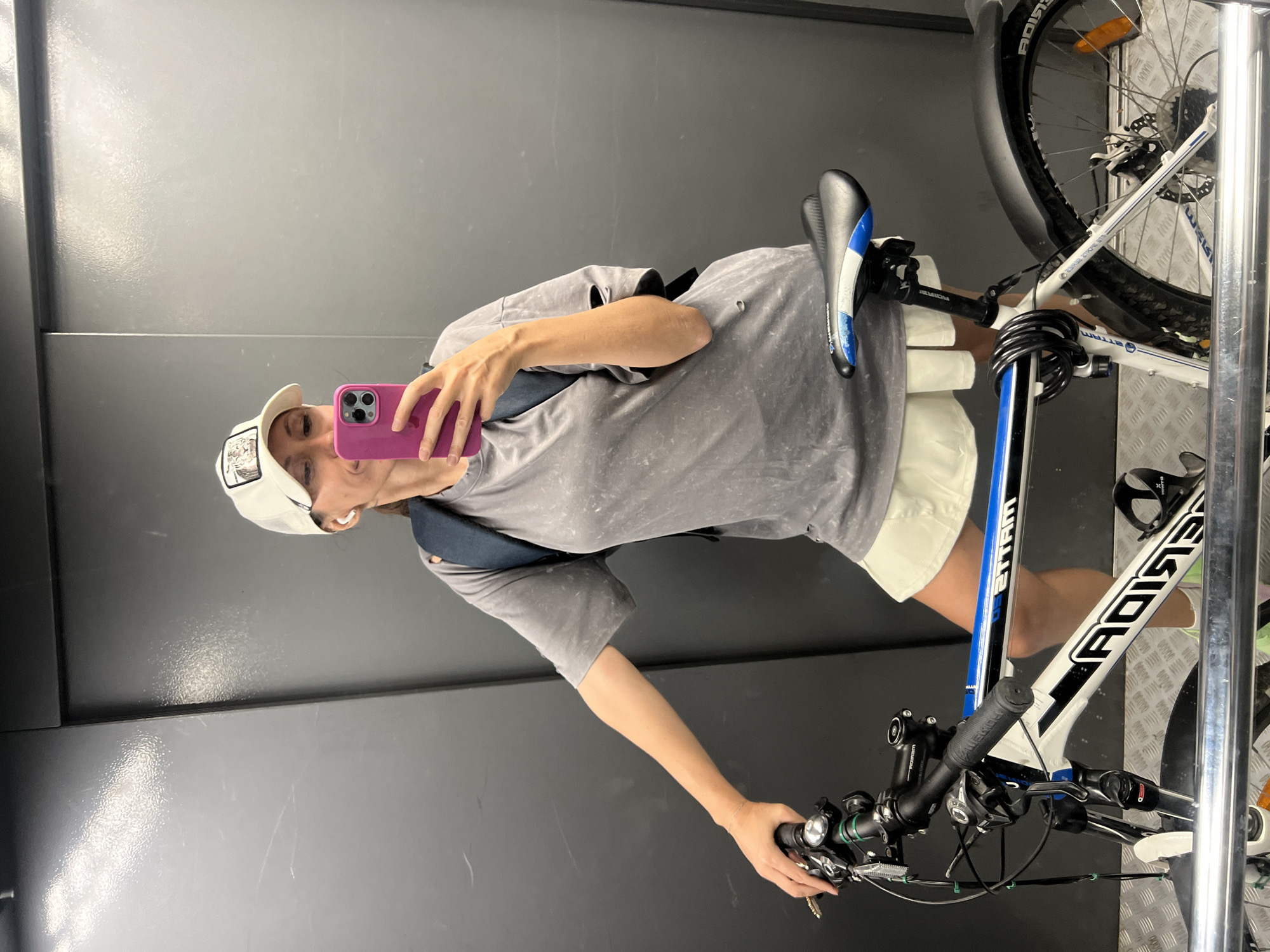 A person is taking a picture of themself with a pink phone while standing with their bicycle in what appears to be an elevator or small, enclosed space with gray walls. The person is wearing a gray t-shirt, white shorts, and a white baseball cap. The bicycle is white with blue accents and has black tires. The scene does not explicitly reveal the location as Moscow, Russia, nor any other city.