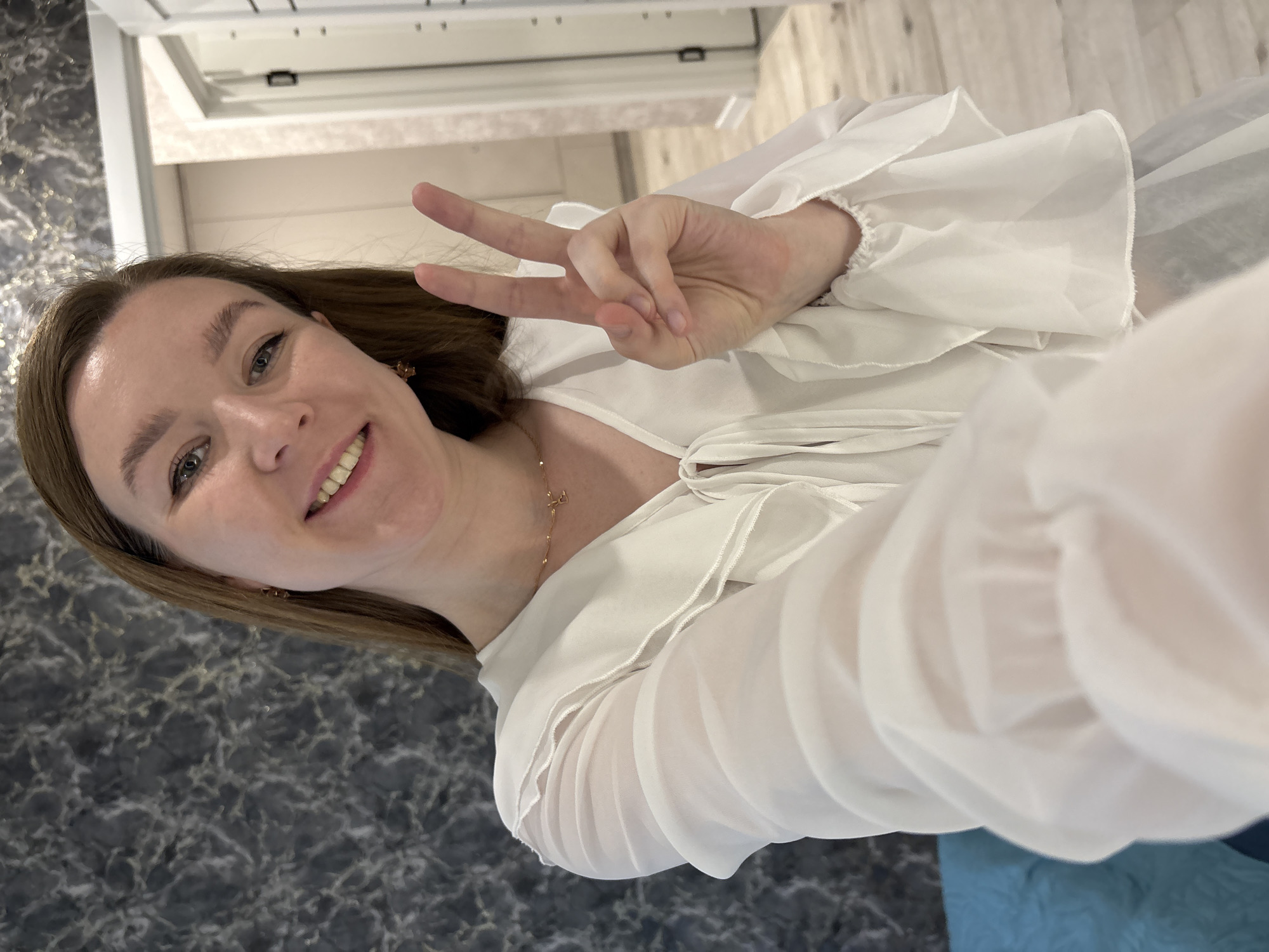 A young woman with light brown hair is captured in a selfie, smiling broadly at the camera while holding up her right hand in a peace or victory sign. She is wearing a white, slightly sheer blouse with ruffled sleeves and a tie-front detail. A delicate gold chain necklace is visible around her neck. The background is tilted due to the selfie angle; to her left (image bottom left due to rotation) is a dark, marble-patterned wall, and to her right (image top right due to rotation) appears to be a lighter wall or door with a floor beyond. The setting is indoors, likely in Izhevsk, Russia. The lighting suggests it is daytime. The woman appears happy and cheerful. There is no visible text in the image.