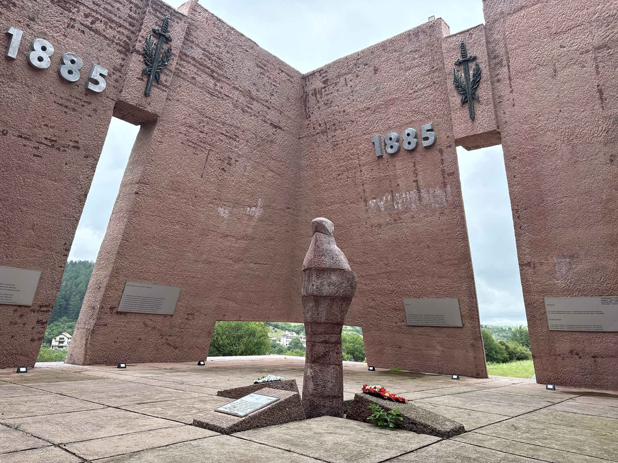 The image depicts the monument to the Battle of Gurgulyat in Gurgulyat, Bulgaria. The monument is made of rough, reddish-brown stone and features large, angular walls with openings. The numbers "1885" are prominently displayed on two of the walls, along with a stylized sword and laurel wreath emblem. In the center of the plaza, a stone sculpture stands, resembling a stylized figure. In front of the sculpture, on the ground, are several stone slabs, some bearing inscriptions and one with a wreath of red flowers and greenery. The plaza is paved with large, square stone tiles. In the background, a green, hilly landscape with trees and some buildings is visible under a cloudy sky. The scene is observed from a low angle, looking upwards towards the monument. There are no people visible in the image.