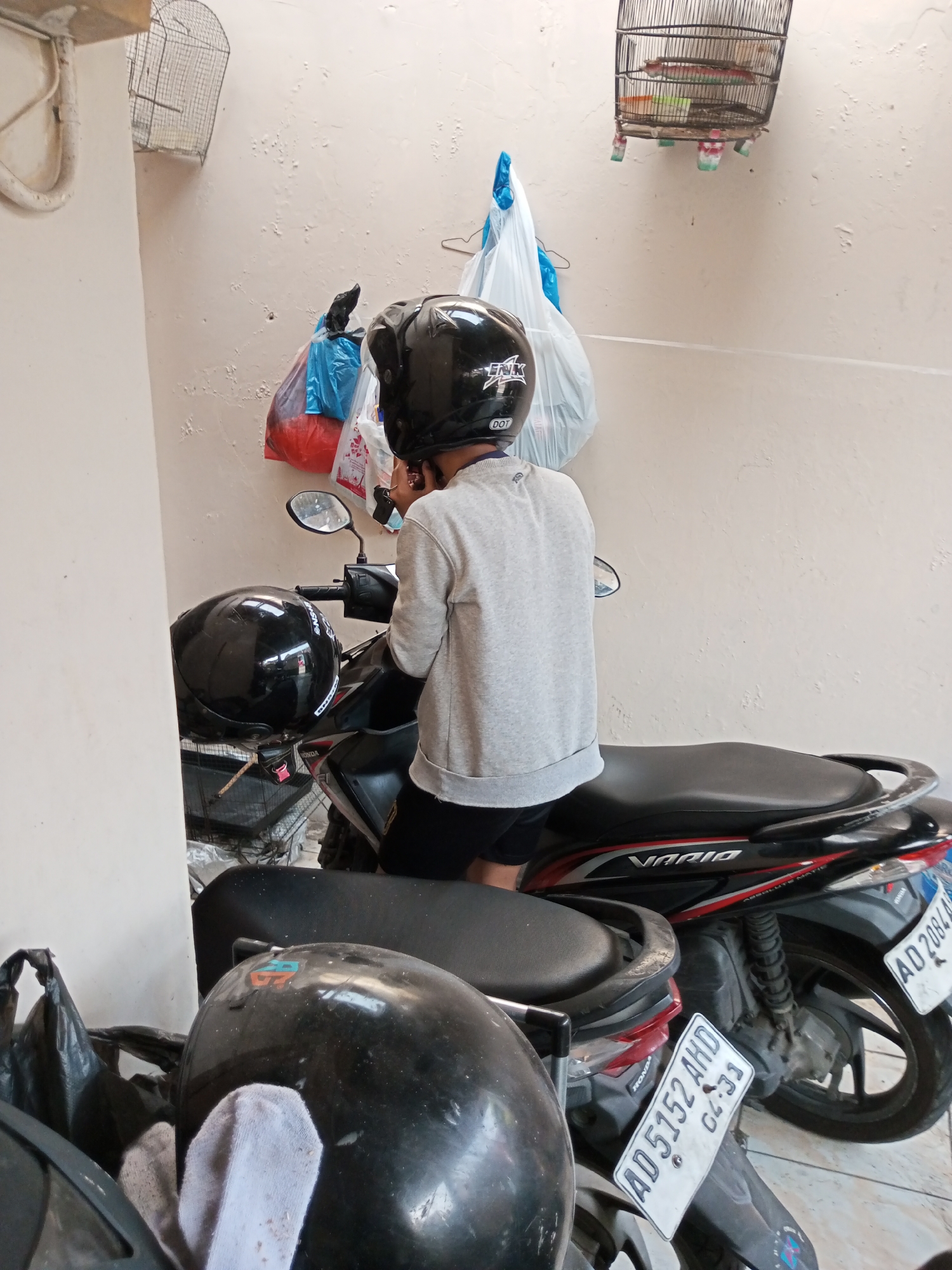The image depicts a person wearing a black helmet and a gray long-sleeved shirt, seated on a black and red Honda Vario scooter. The scooter is parked next to another scooter, also with a black helmet placed on its seat. The scene appears to be outdoors, with a textured, light-colored wall in the background. Various items are hanging on the wall, including plastic bags and a birdcage. The ground is tiled. The license plate of the visible scooter reads "AD 2084 AD". The presence of the Honda Vario scooter and the license plate format suggests the location is likely in Indonesia, possibly in or near Banyudono, based on the license plate prefix which is often associated with Central Java.