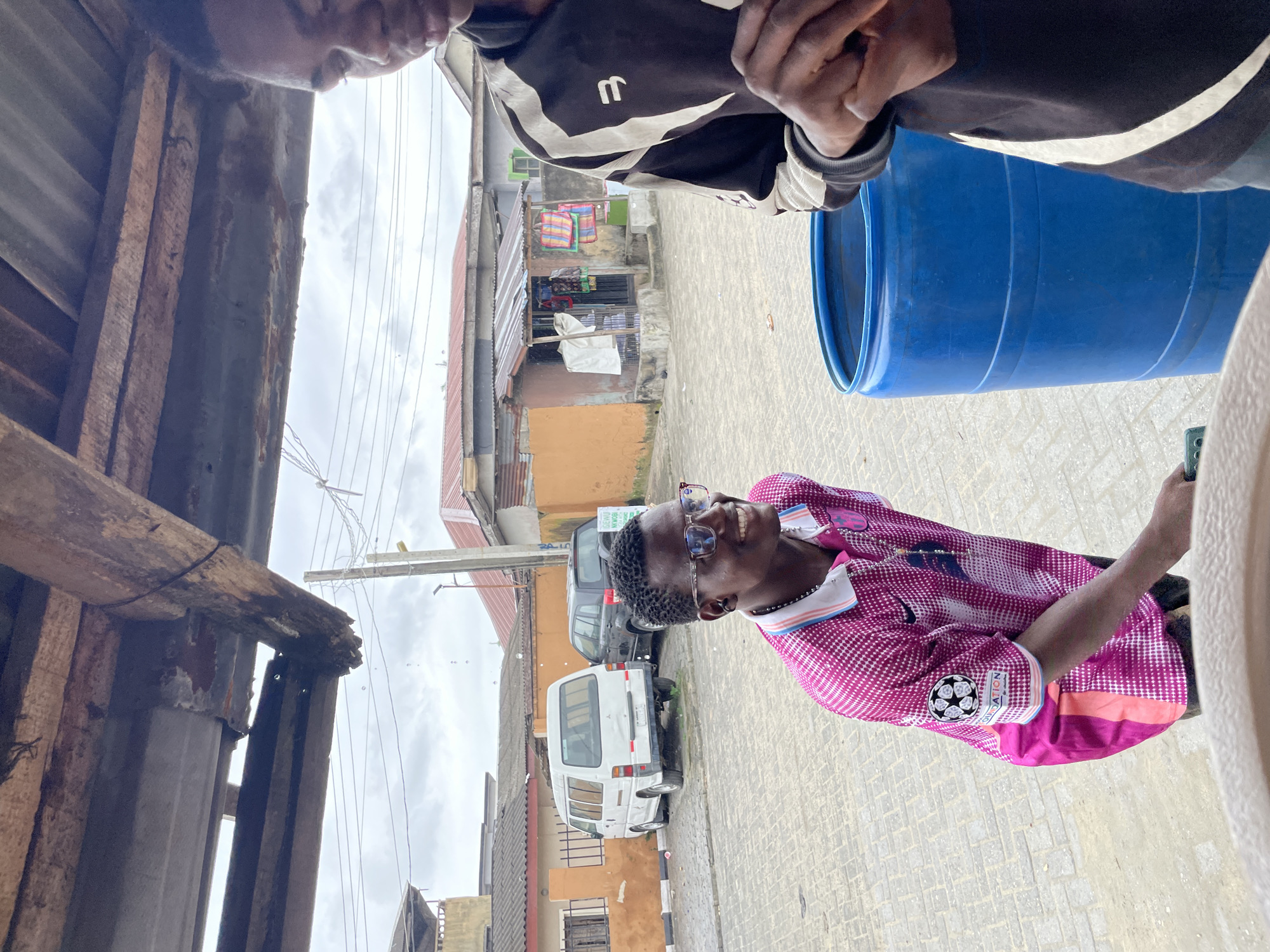 In Ado, Nigeria, an outdoor scene unfolds under a weathered, rustic wooden overhang. Two young men are present. The man in the foreground on the left is wearing a pink shirt with a white and blue collar. He has glasses and is smiling. The second man, on the right, is wearing a black and white jacket and is partially obscured by the frame. In the background, a white van and a dark SUV are parked. Behind them, a street lined with buildings and small shops is visible. A large blue barrel is situated between the two men. The ground is paved with interlocking bricks, and the sky is overcast.