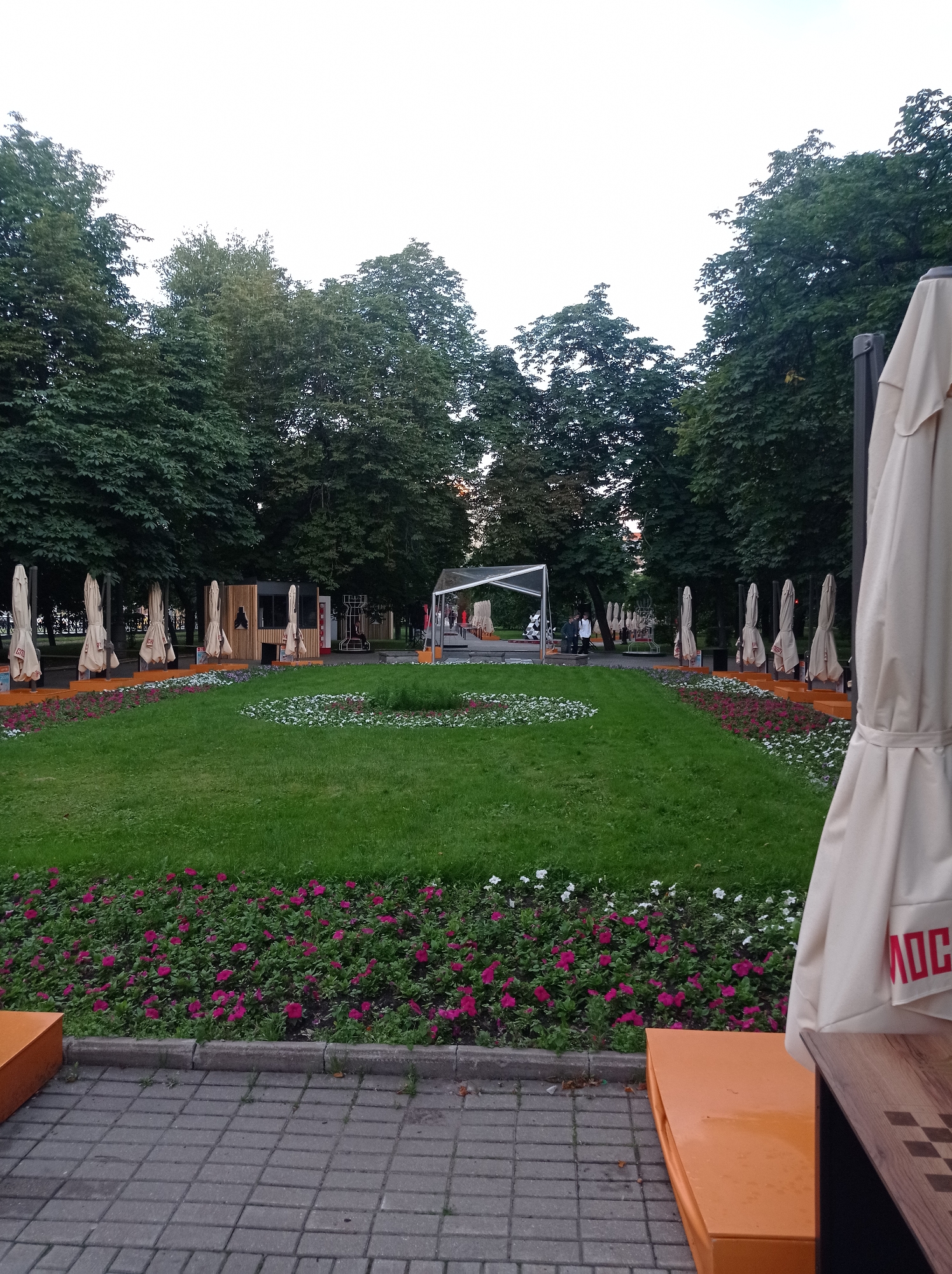 The image depicts an outdoor public space, likely a park or square, in Moscow, Russia. In the foreground, a beige umbrella is partially visible on the right, with red lettering that appears to spell "MOC" on its fabric. To the left of the umbrella, an orange bench or seating area is situated. Beyond this, a paved walkway made of gray rectangular tiles stretches into the distance.
The main area of the image features a well-maintained lawn with flowerbeds along its edges. The flowerbeds are planted with a mix of pink and white flowers. In the mid-ground, a temporary structure, possibly a stage or an informational booth, is set up with a white canopy. A few people are visible near this structure.
Further back, a row of closed umbrellas, all uniformly beige, stands along an elevated platform. These are likely outdoor seating umbrellas for a cafe or restaurant that is not actively in use. The background is dominated by lush green trees with dense foliage. The sky above is overcast and light in color. The overall impression is of a quiet, possibly early evening or daytime scene in a recreational area of the city.