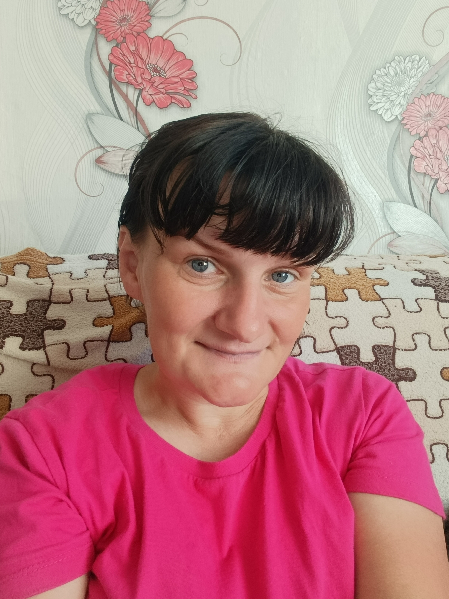 A woman with short dark hair and blue eyes is seated in front of a wall with a floral pattern. She is wearing a bright pink t-shirt. Behind her is a patterned cushion or blanket that resembles puzzle pieces. The location in the city of Фоки, Russia is unconfirmed.