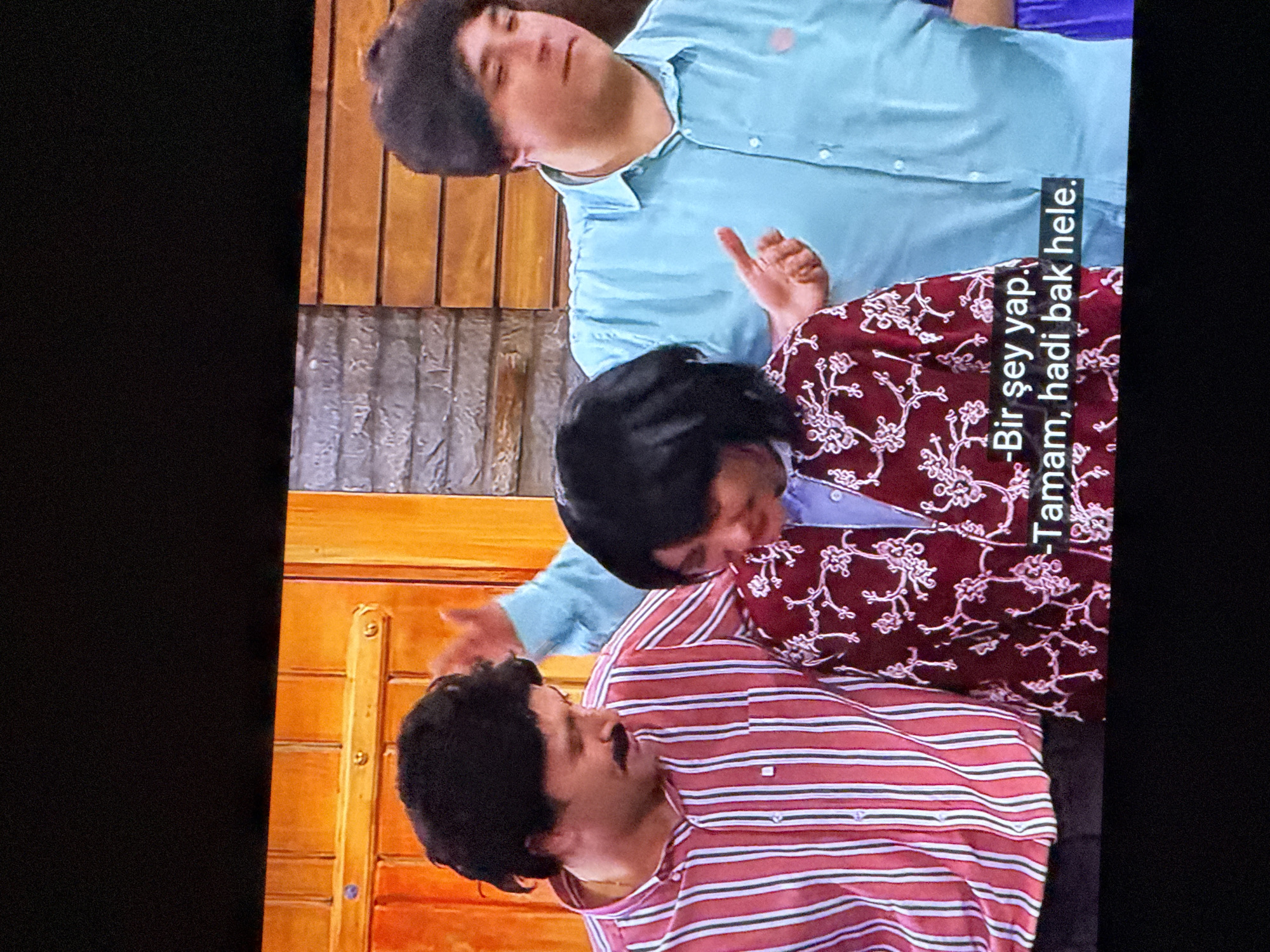 The image shows three people standing in front of an orange door and a wooden wall. The person on the left is a man with dark hair and a mustache, wearing a red and white striped shirt. He is looking down. In the center is a person with dark, short hair, wearing a dark red jacket with a white floral pattern over a light blue shirt. This person is also looking down. To the right is a person with dark, curly hair, wearing a light blue button-down shirt. This person is looking to their right and has their right hand raised. Below the two people in the center and right are subtitles that read: "-Bir şey yap. -Tamam, hadi bak hele." The scene appears to be indoors, possibly on a set. No specific location in Waregem, Belgium can be identified from this image.