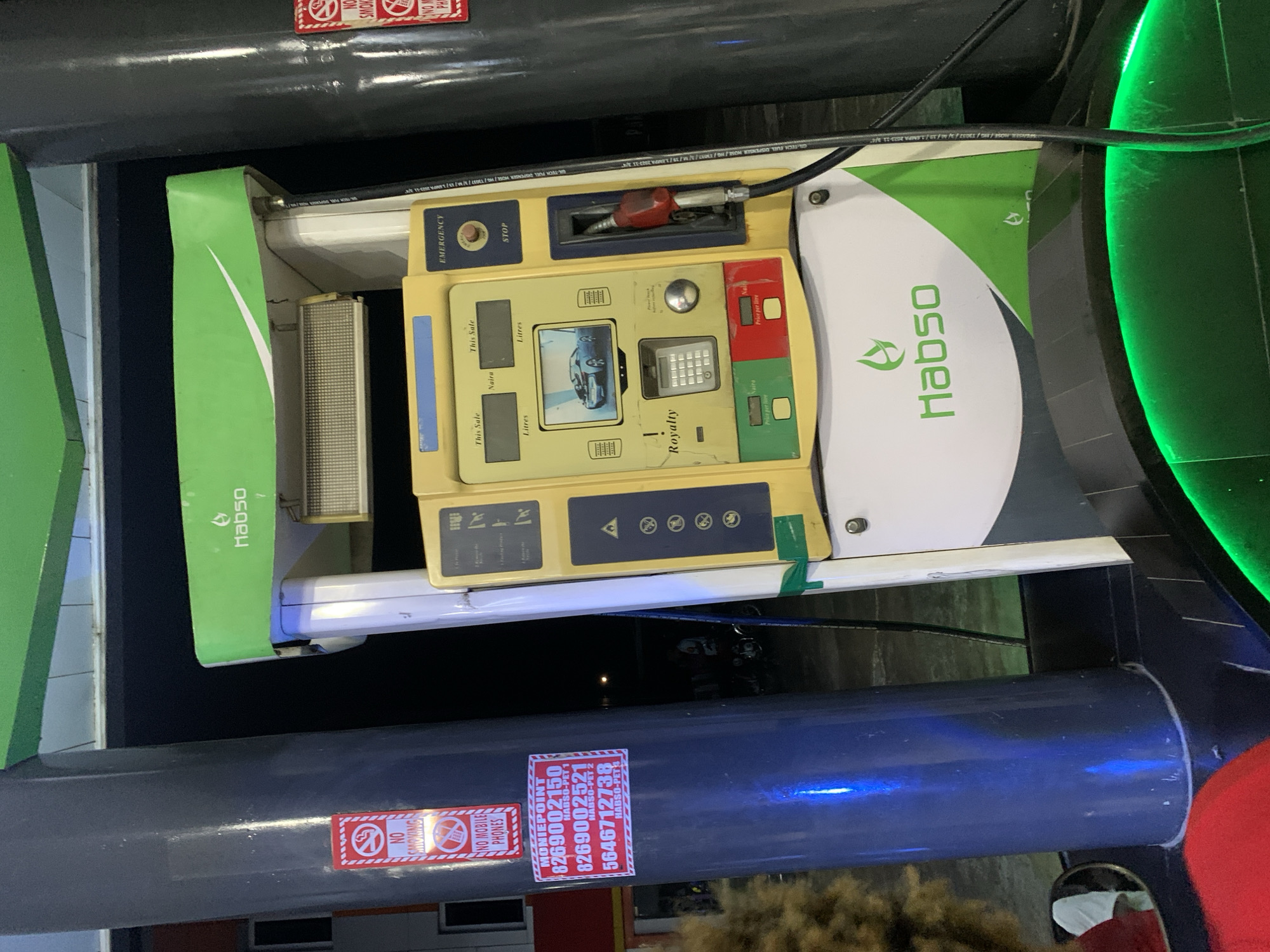 The image shows a Habso-branded fuel dispensing station in Lokoja, Nigeria. The pump, painted in green and white, features an emergency stop button and a digital display showing "This Sale" information in Naira and Litres. The pump has a keypad labeled "Royalty" and other buttons beneath the screen. Nozzles are mounted beside the pump, with a red handle. The fuel station infrastructure includes a dark grey cylindrical structure with "NO SMOKING" and "NO MOBILE PHONES" warning signs. A sticker bearing the text "MONIEPOINT" followed by several phone numbers and "Habso-PET" identifiers is visible on the structure. Bright green neon lighting fixtures appear nearby. The scene is captured at night with limited visible ambient light.