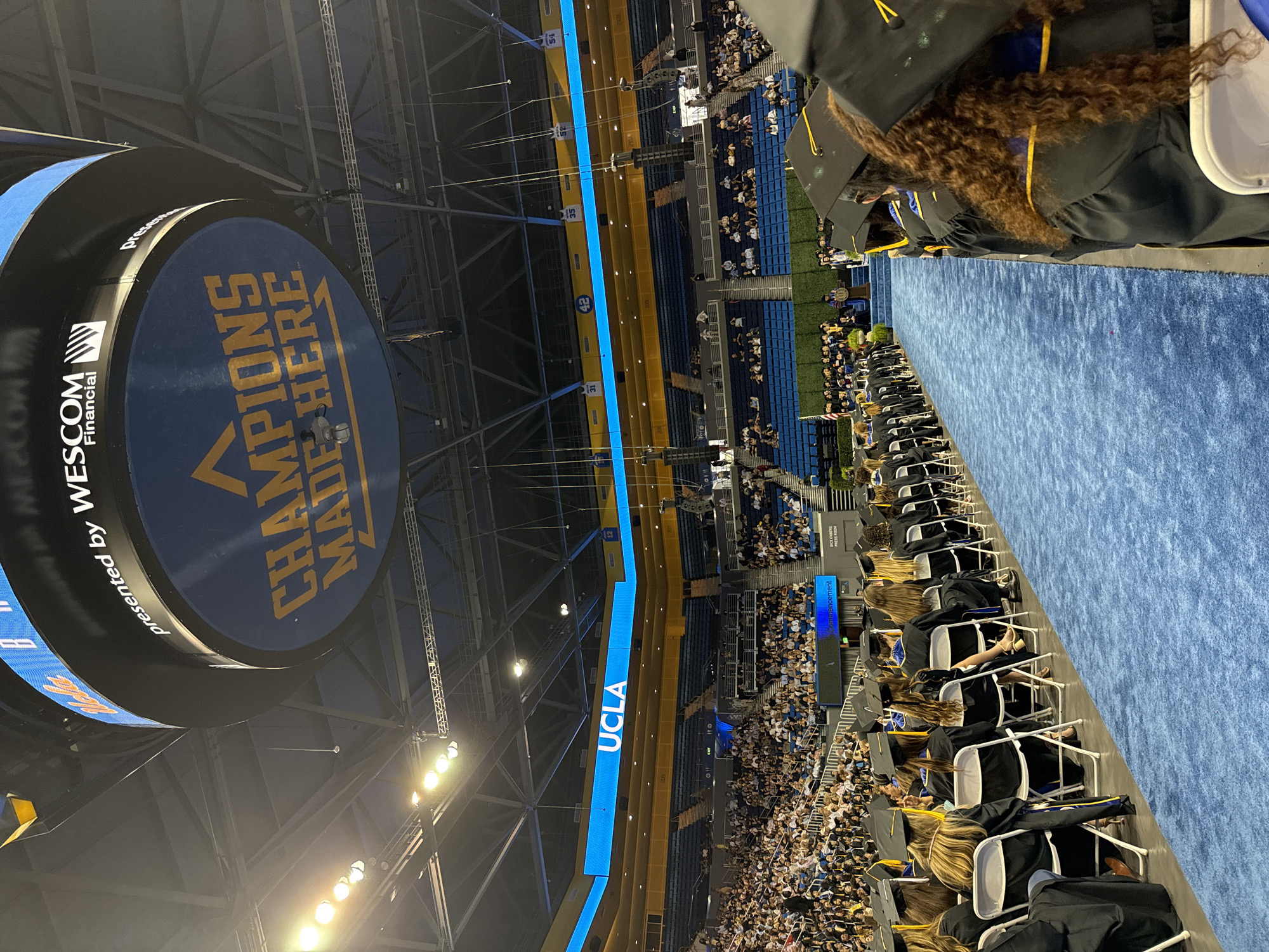 The image depicts a graduation ceremony at the Pauley Pavilion at UCLA in Los Angeles, California. A large number of graduates, dressed in black gowns and caps, are seated in rows on a blue carpeted aisle, facing a stage area. The background shows tiered seating filled with spectators. Above the seating, a large digital scoreboard displays "UCLA" in bright blue letters. Another large circular sign, suspended from the ceiling, reads "CHAMPIONS MADE HERE" in gold lettering on a blue background, with "presented by WESCOM Financial" visible above it. The arena is illuminated by stadium lighting.