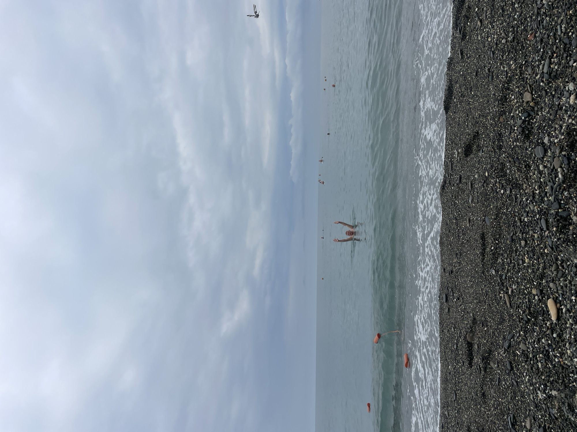 A single person is visible in the gray ocean, waist-deep, with their arms raised. The person is in the middle of the frame, facing the camera. Several orange buoys are scattered in the water. The beach in the foreground is covered in dark pebbles. The sky is overcast with gray clouds. The scene is set on a pebble beach in Sochi, Russia.