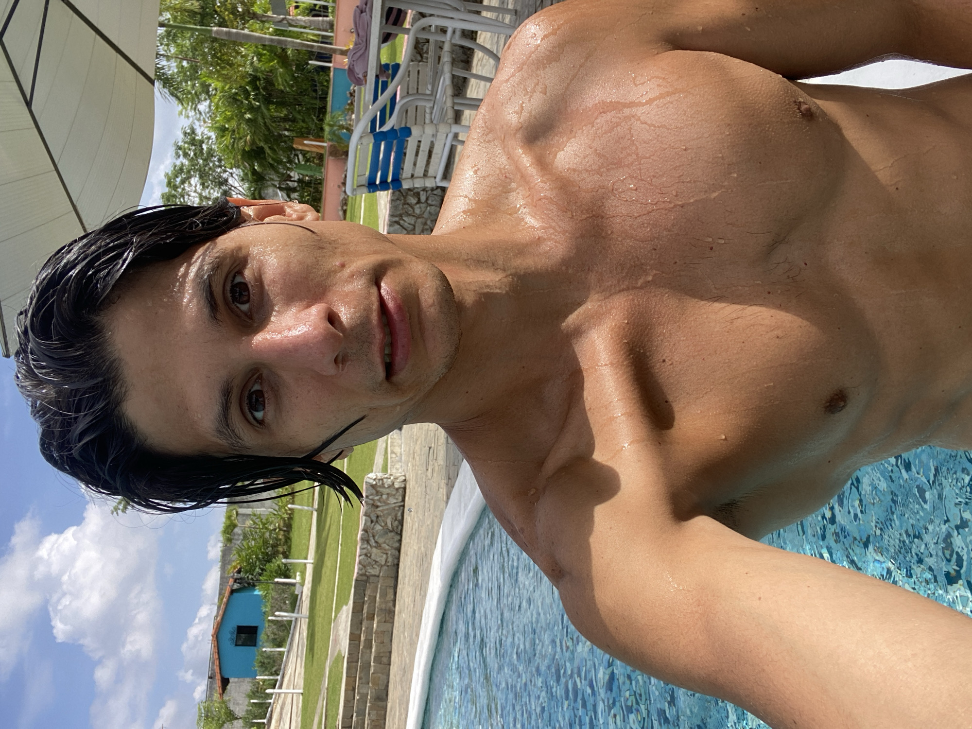 The image is a selfie taken by a man, likely in his 20s or 30s, with dark, wet hair, looking directly into the camera. His bare upper torso displays muscle definition and is covered in visible water droplets, suggesting he has recently been in a pool. He has a neutral expression, with a slight hint of a smile.
The scene is an outdoor setting at a swimming pool, under bright, sunny conditions. The man is positioned at the edge of a pool with blue mosaic tiling, which is visible in the lower and right parts of the frame.
In the background, a large white, angled canopy or umbrella structure provides shade on the left. Beyond that, white outdoor chairs and tables are arranged, indicating a poolside lounge area. Further back, lush green trees and foliage are present under a clear blue sky with scattered white clouds. To the right, a distinctive small, bright blue building with a dark window stands on a manicured grassy lawn, adjacent to what appears to be stone steps or terraced landscaping. The strong sunlight casts clear shadows and highlights the wetness on the man's skin and hair. The setting suggests a leisurely, vacation-like atmosphere, consistent with Vallecito, Venezuela. No visible text is present in the image.
