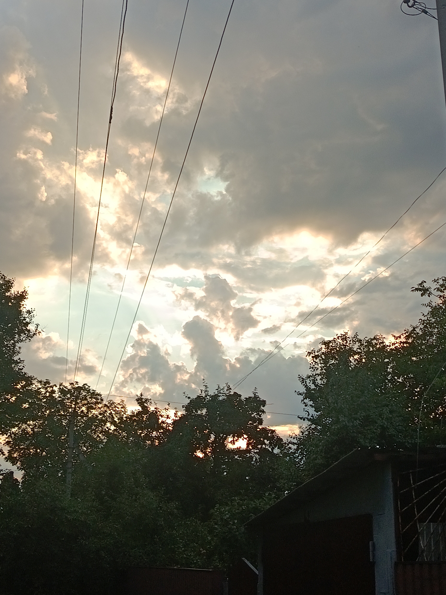 The image depicts an outdoor scene in Smila, Ukraine, during the evening. The sky is filled with dramatic, partly cloudy conditions, with sunlight breaking through the clouds in several areas, creating a luminous effect. Multiple utility wires stretch across the frame from left to right, diagonally dividing the sky. Silhouetted against the bright sky are the tops of lush green trees and foliage, suggesting a natural or semi-rural setting within the city. In the lower right corner, a portion of a building with a sloped roof and a metallic grate is visible, indicating a residential or utility area. The overall mood is somewhat somber yet beautiful due to the interplay of light and shadow. There are no people or discernible actions taking place in the image.
