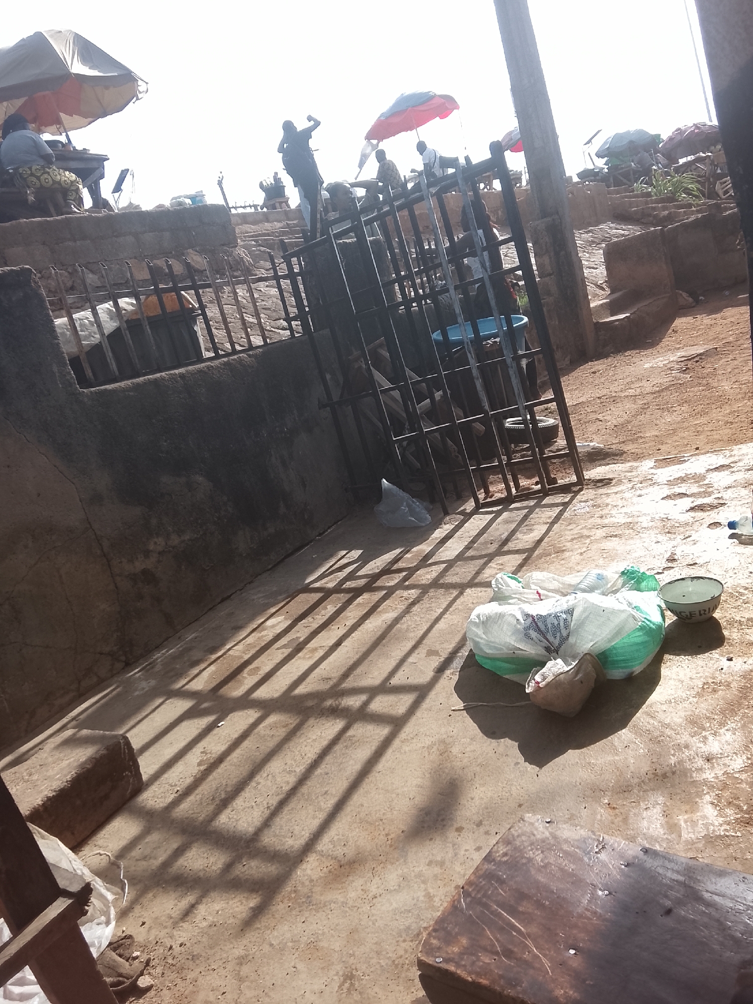 The image depicts an outdoor scene in Gwong, Nigeria. A collection of plastic bags and a metal bowl sit on a concrete surface, partially shadowed by a metal fence. Above and beyond the fence, there is a raised area with people, stalls, and umbrellas indicating a marketplace. One man appears to be raising his arm. The general environment is dusty and shows signs of wear and tear.