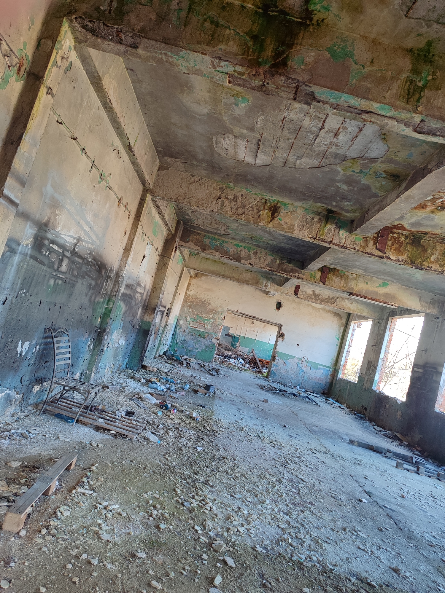 The image shows the interior of an abandoned building. The location is presumed to be Burgas, Bulgaria. The scene is characterized by significant decay, with crumbling concrete walls and ceiling. Paint is peeling and flaking. Debris is scattered across the floor, including wood fragments, trash, and rubble. A metal chair-like object sits against one wall. Windows are visible, revealing glimpses of an exterior scene. The lighting is diffused, suggesting natural illumination through the openings in the building's structure. There is no indication of human presence in the immediate view.