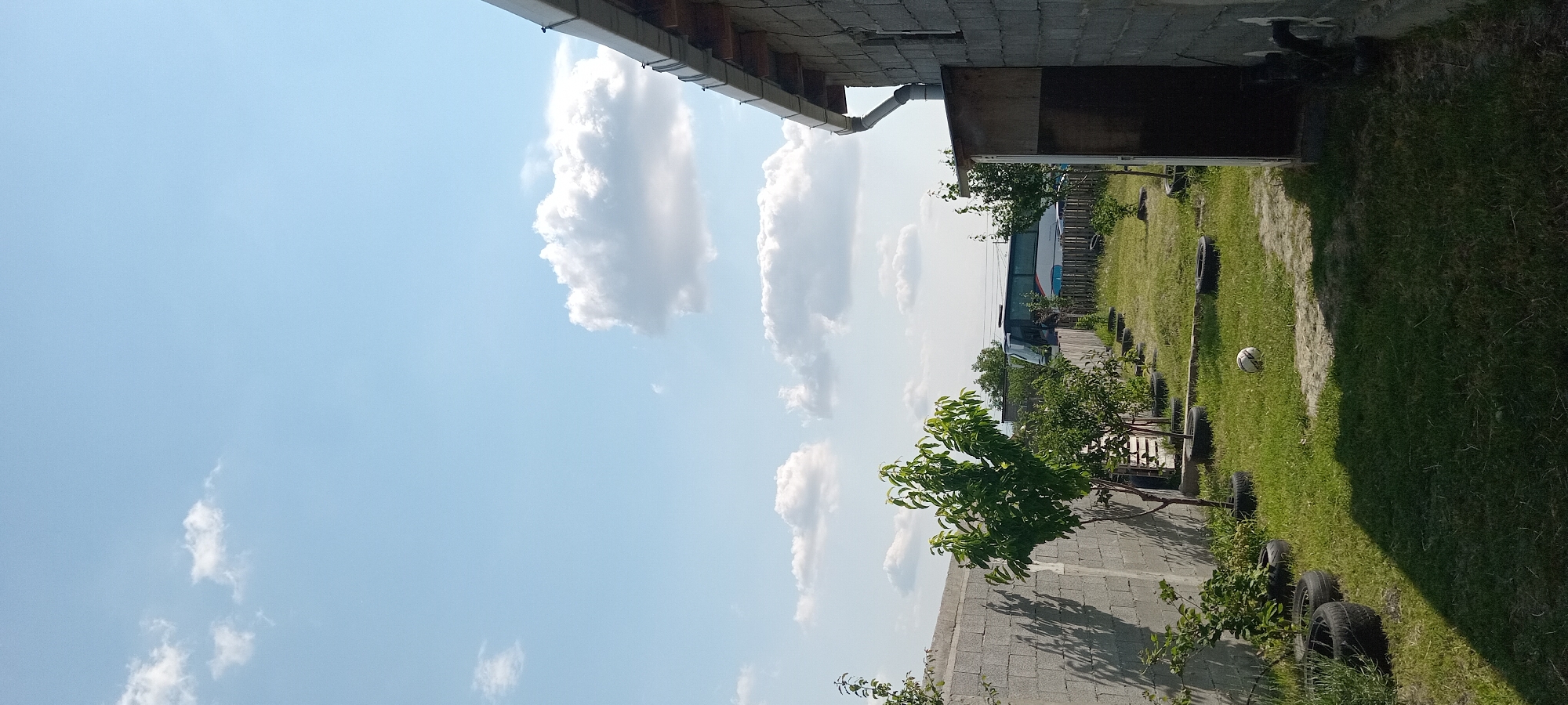 The image depicts an outdoor scene on a bright, sunny day. The sky is a clear, pale blue, dotted with fluffy white clouds. In the foreground, a section of a building with a concrete block wall and a metal roof overhang is visible. A downspout extends from the roof.
To the right of the building, a grassy area stretches out, lined with what appear to be old tires placed vertically. A soccer ball rests on the grass near the tires. Beyond the grassy area, trees and bushes with green foliage are scattered throughout the scene. In the background, a large white bus with a blue stripe is parked. The overall setting appears to be a yard or an open area adjacent to a building, possibly a rural or suburban location. The lighting suggests it is daytime. There is no visible text in the image.