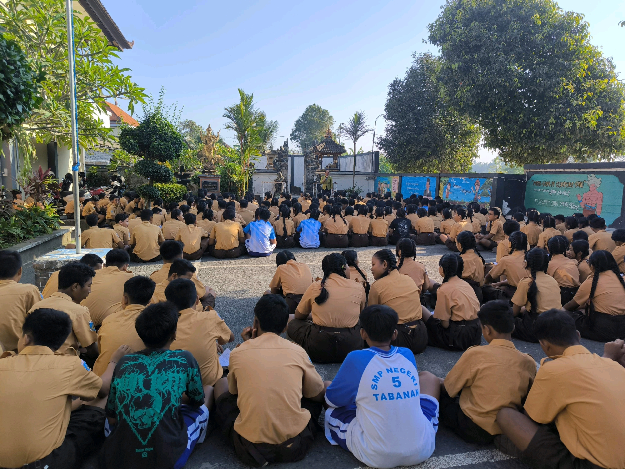 In Tabanan, Indonesia, a large group of students, primarily male and dressed in brown school uniforms, are seated on the ground in what appears to be an outdoor assembly area. The students are arranged in concentric circles, facing inward. In the background, a man in a light-colored jacket stands before a stage or platform, seemingly addressing the students. Behind the man is a wall decorated with colorful murals. The environment includes lush trees and vegetation, suggesting a tropical setting. The sky is clear and blue, indicating a sunny day. The students' postures and focus suggest they are attentively listening to the speaker.