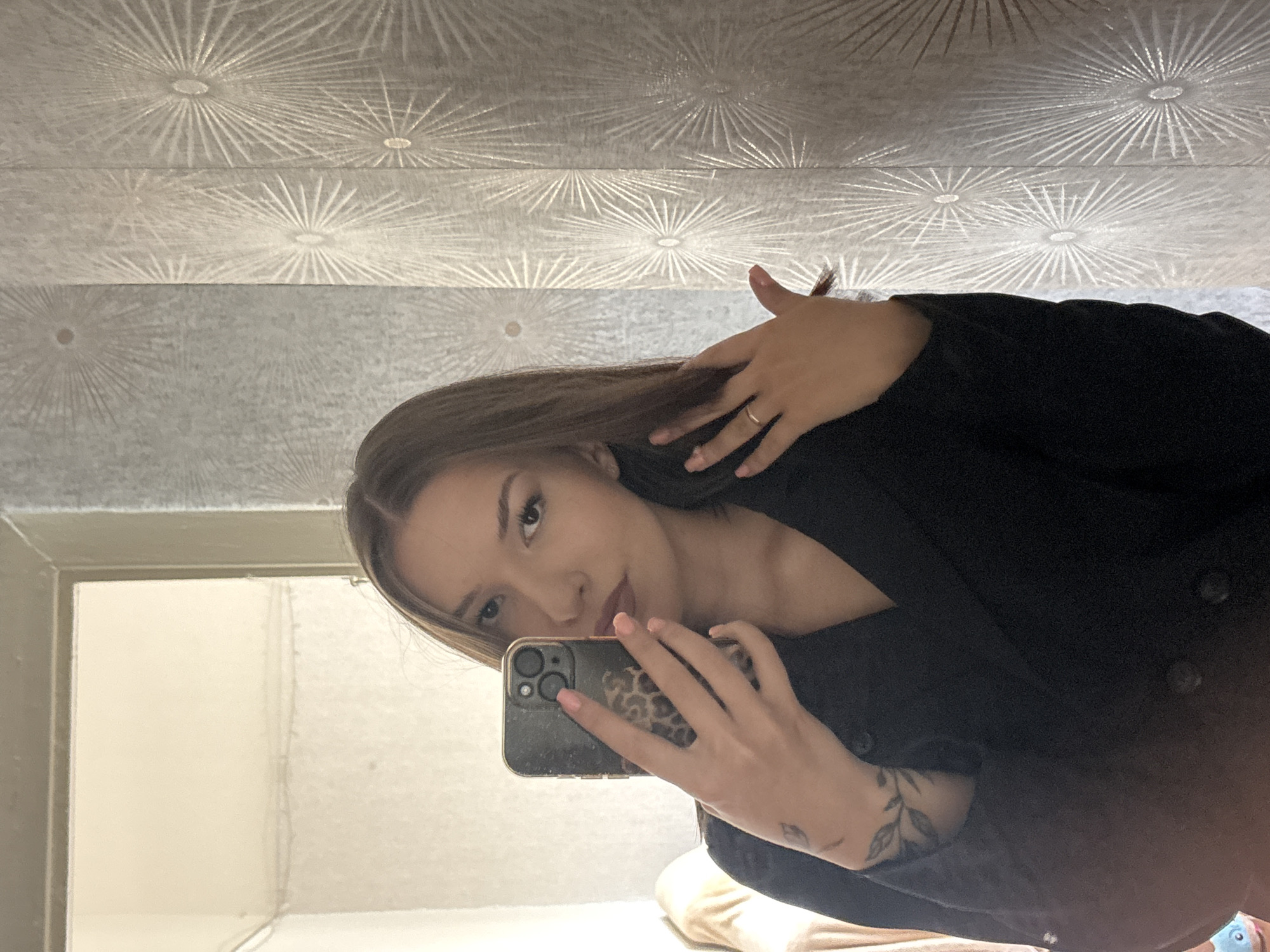 A young woman with long brown hair takes a selfie in a room with patterned wallpaper. She is wearing a black jacket and has a tattoo of a branch on her left forearm. Her right hand is touching her hair, and she is holding a smartphone in her left hand. The wallpaper has a starburst pattern in a silver tone. The image is taken indoors, and there is a mirror reflecting the scene. There is no visible indication of the location being in the city of Inskoy, Russia.