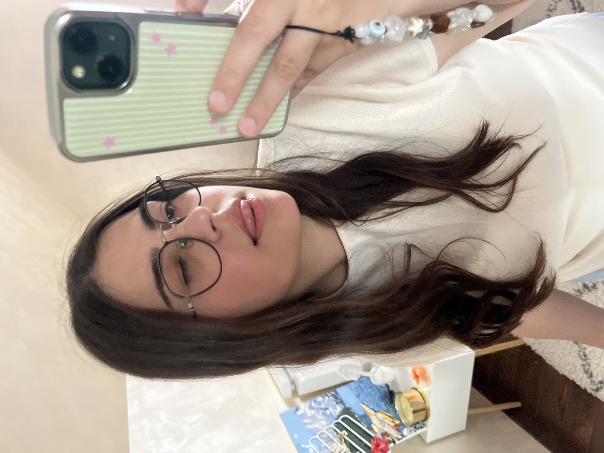 A young woman with long, dark brown hair, wearing glasses and a white t-shirt, is taking a selfie with her phone. She is winking and sticking her tongue out. Her phone has a light green case with pink stars. A beaded charm hangs from the phone. In the background, there is a white nightstand with a painting of mountains and water, a golden candle, and other small objects. The room appears to be a bedroom. The image is taken indoors, possibly in an apartment in Koroviia, Ukraine.