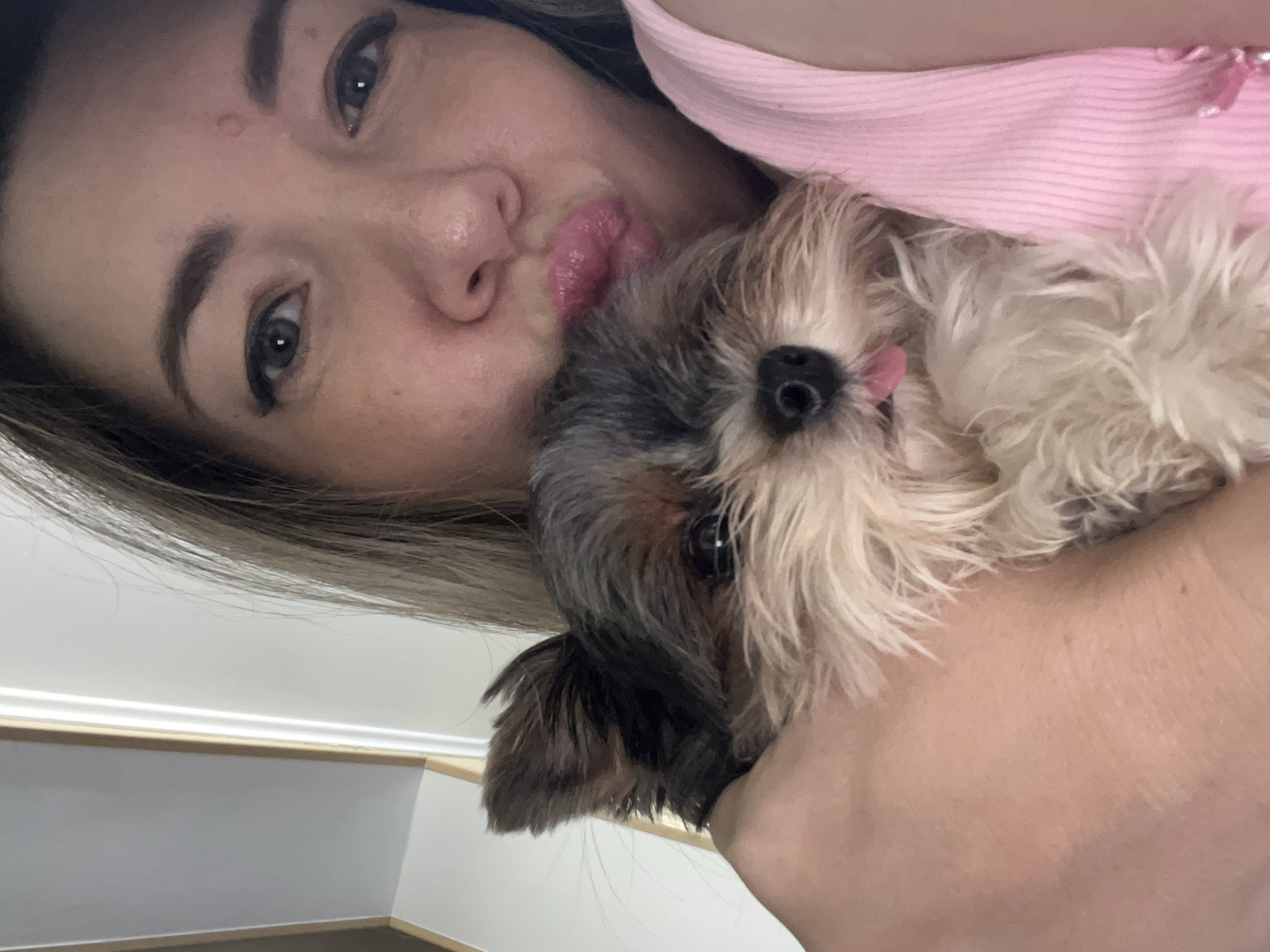 In this close-up, indoor selfie, a woman with blonde hair and makeup is holding a small dog. The woman's face is partially obscured as she puckers her lips, leaning in towards the dog. The dog, a Yorkshire Terrier, has its tongue sticking out slightly. The woman is wearing a pink tank top. The background is indistinct, appearing to be a wall with some vertical trim. There is no indication of the city of Sochi, Russia in the image.