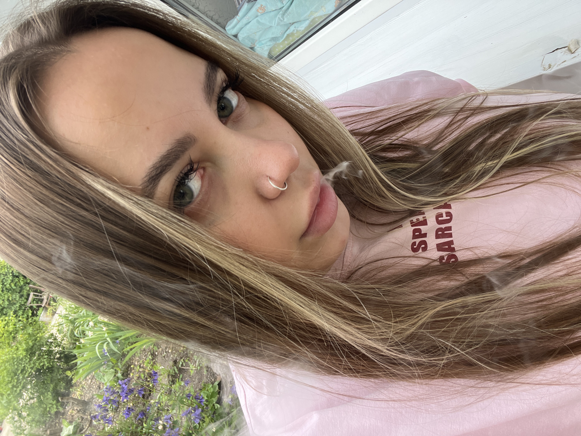A young woman with long, highlighted brown hair and green eyes is depicted from a close, low-angle perspective. She has a silver nose ring and is exhaling smoke. She is wearing a pink shirt with writing on it. The background consists of an exterior view of green foliage, including some purple flowers, and a window frame. The image appears to be taken indoors, near a window. The city of Novopokrovskaya, Russia, cannot be determined from the visual information provided.