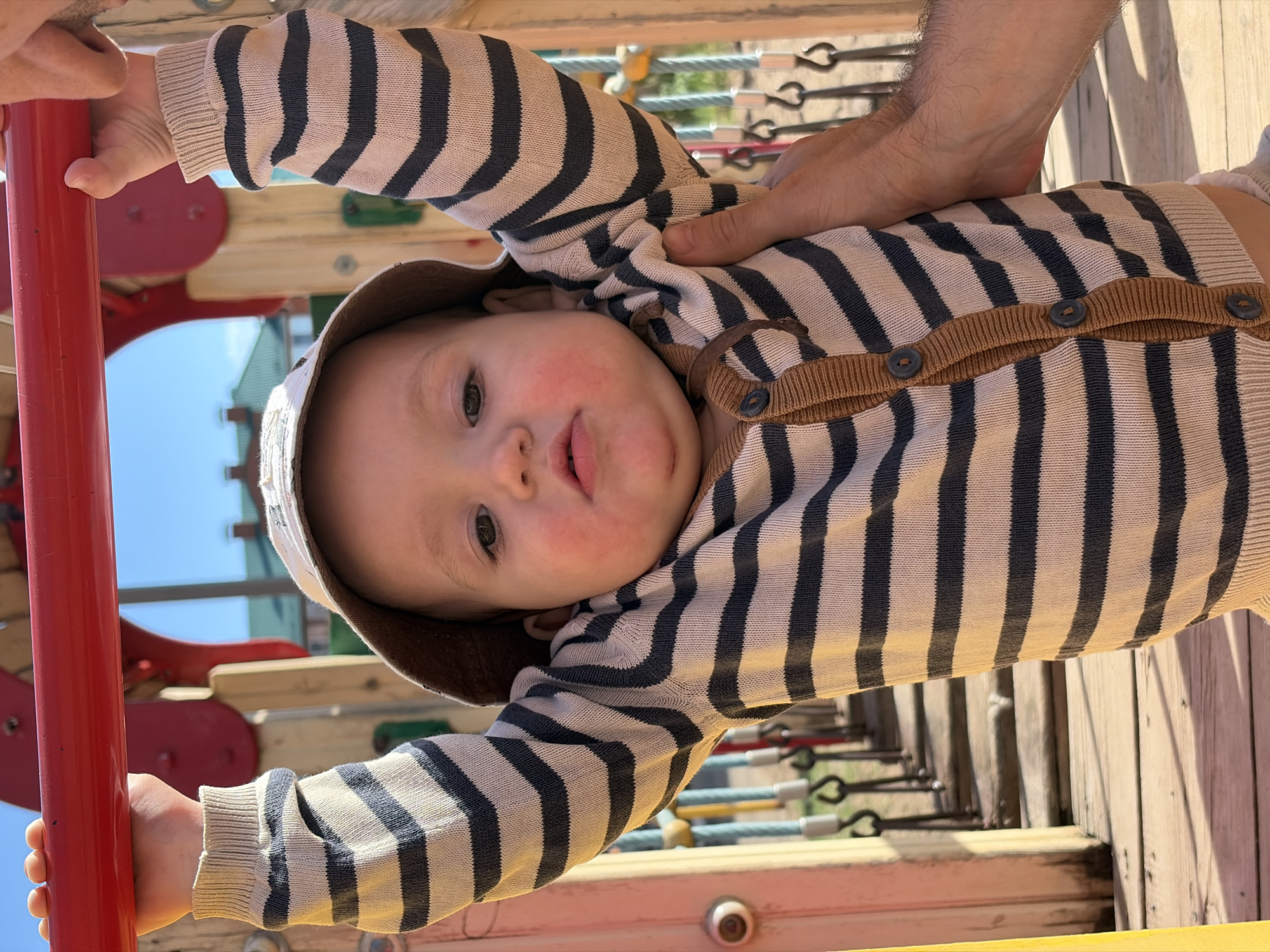 A baby in a striped cardigan and a hat is on a playground. The baby is holding onto a red pole with their left hand. A person's hand is near the baby's chest. The playground has wooden and metal structures. The location is likely in the city of Chaikovsky, Russia.