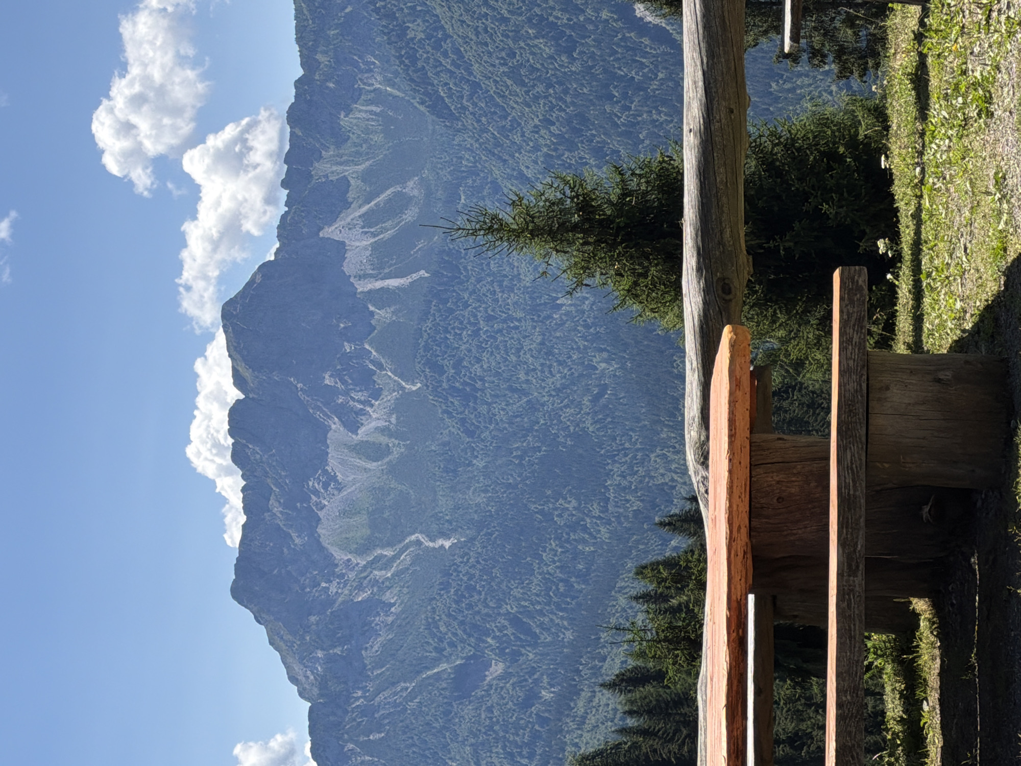 A close-up view of a wooden picnic table and benches is in the foreground of an expansive mountain landscape. The table and benches are constructed from rough-hewn logs and planks, showing natural wood grain and color. The backdrop features a dense, dark green forest covering the slopes of a large mountain range. The mountain peaks are rugged and rocky, with visible ravines and lighter-colored streaks, suggesting scree or exposed rock faces. The sky above is a clear, bright blue with scattered, fluffy white clouds. The scene is set outdoors, with the picnic area on a grassy, sun-dappled hillside.