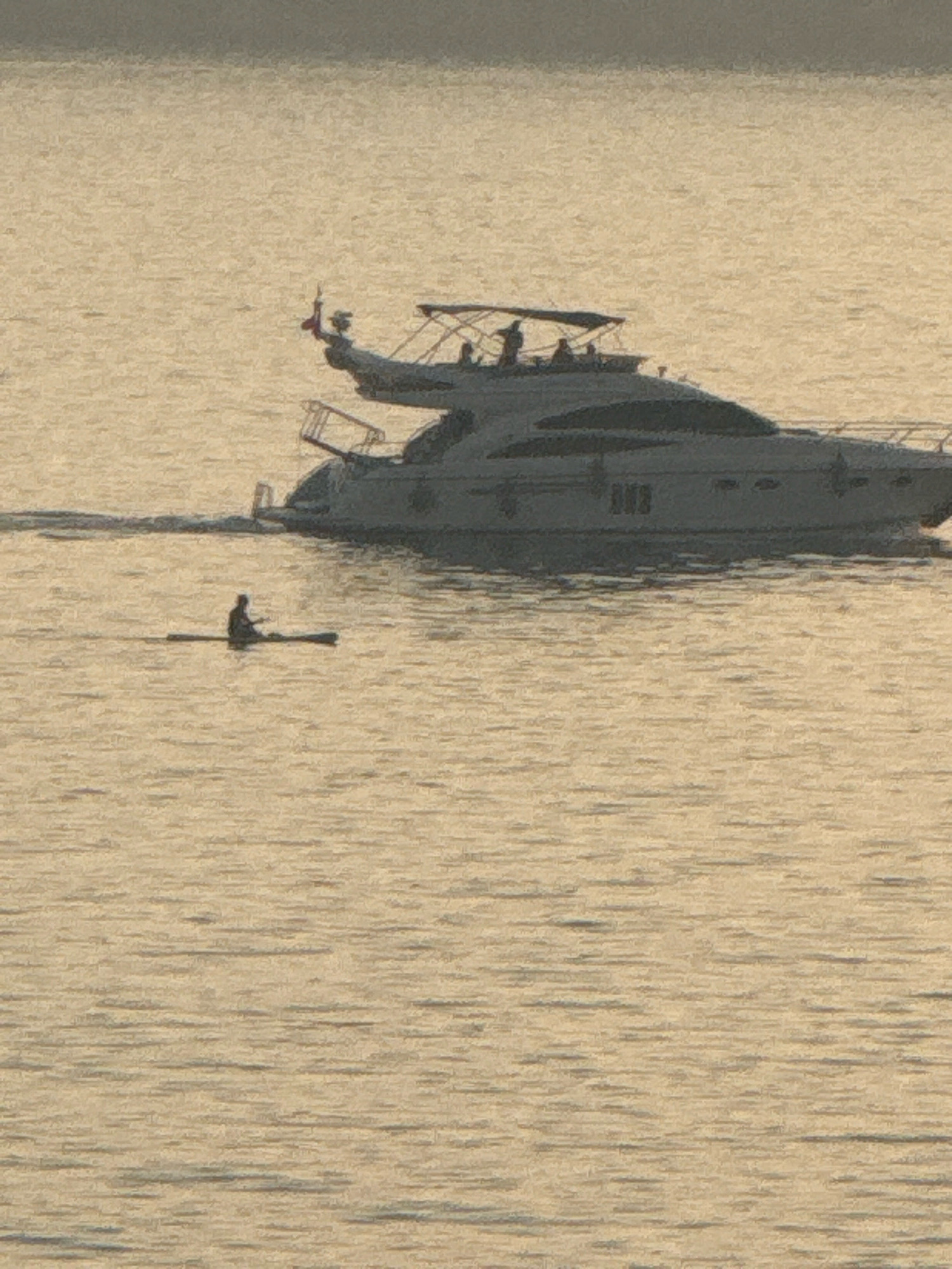 In Kamenovo, Montenegro, on a calm body of water reflecting a muted, possibly hazy sky, a large, modern yacht is observed at anchor. The yacht, with its sleek, dark hull and multiple levels, is a prominent feature. On its deck, several figures are visible, though their actions are indistinct. In the foreground, a solitary individual is seated on a stand-up paddleboard, appearing to be paddling. The water is a light, sandy color, with subtle ripples suggesting minimal movement. The overall atmosphere is serene and still.