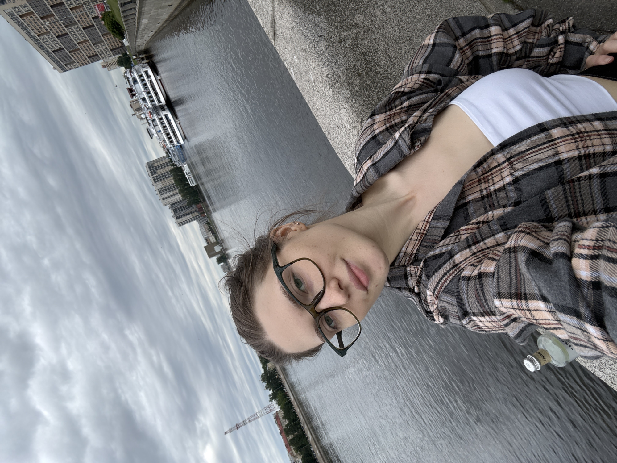 A young woman wearing glasses and a plaid shirt is taking a selfie outdoors. She is standing near a body of water, likely a river, with a cityscape in the background. Several buildings and what appear to be cruise ships are visible across the water. The sky is overcast with clouds. There is a small bottle on the ledge next to her. The location is identified as Shushary, Russia.