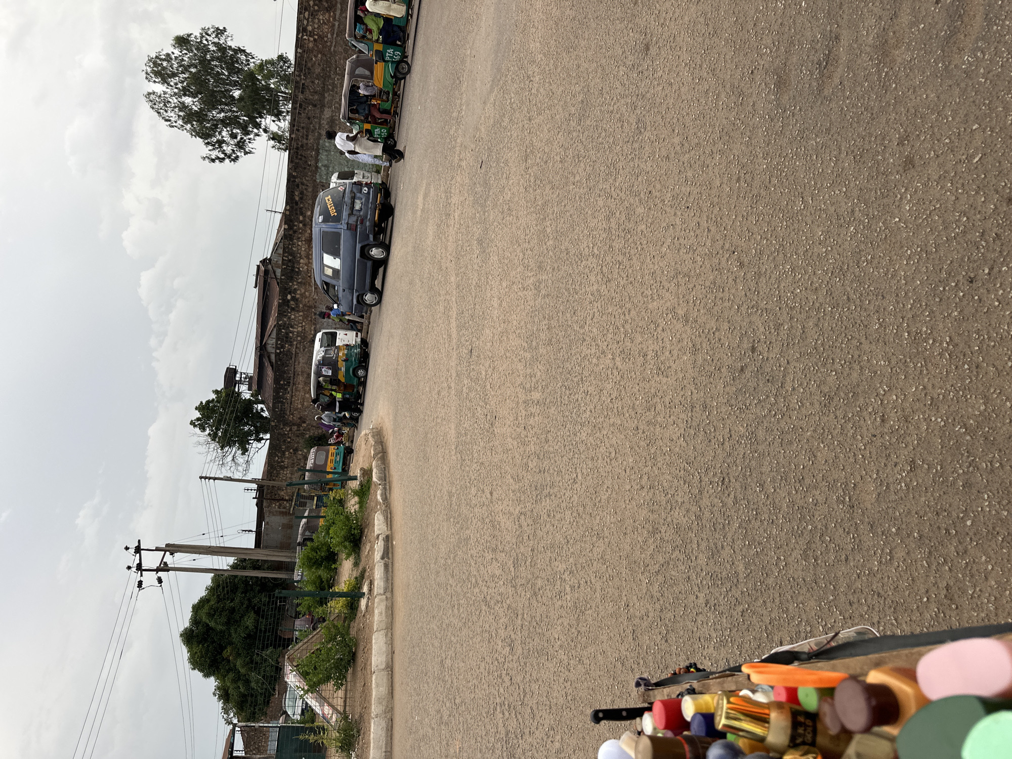 This is a street-level, medium-angle, medium shot of a dusty road with a small market stall in the foreground on the left. The stall displays an assortment of colorful bottles, some with metallic gold labels. In the background, a grey, cloudy sky looms over a street lined with parked vehicles, including several auto-rickshaws, a dark blue van with the word "JUSTICE" emblazoned in yellow on the rear door, and another white van. A few people are visible near the vehicles, some appear to be passengers and others may be vendors. The scene is set in Bukuru, Nigeria, with a stone wall and some trees forming the backdrop. The road is paved with asphalt and shows signs of wear, with scattered gravel. The overall impression is of a typical, bustling day in a Nigerian city.