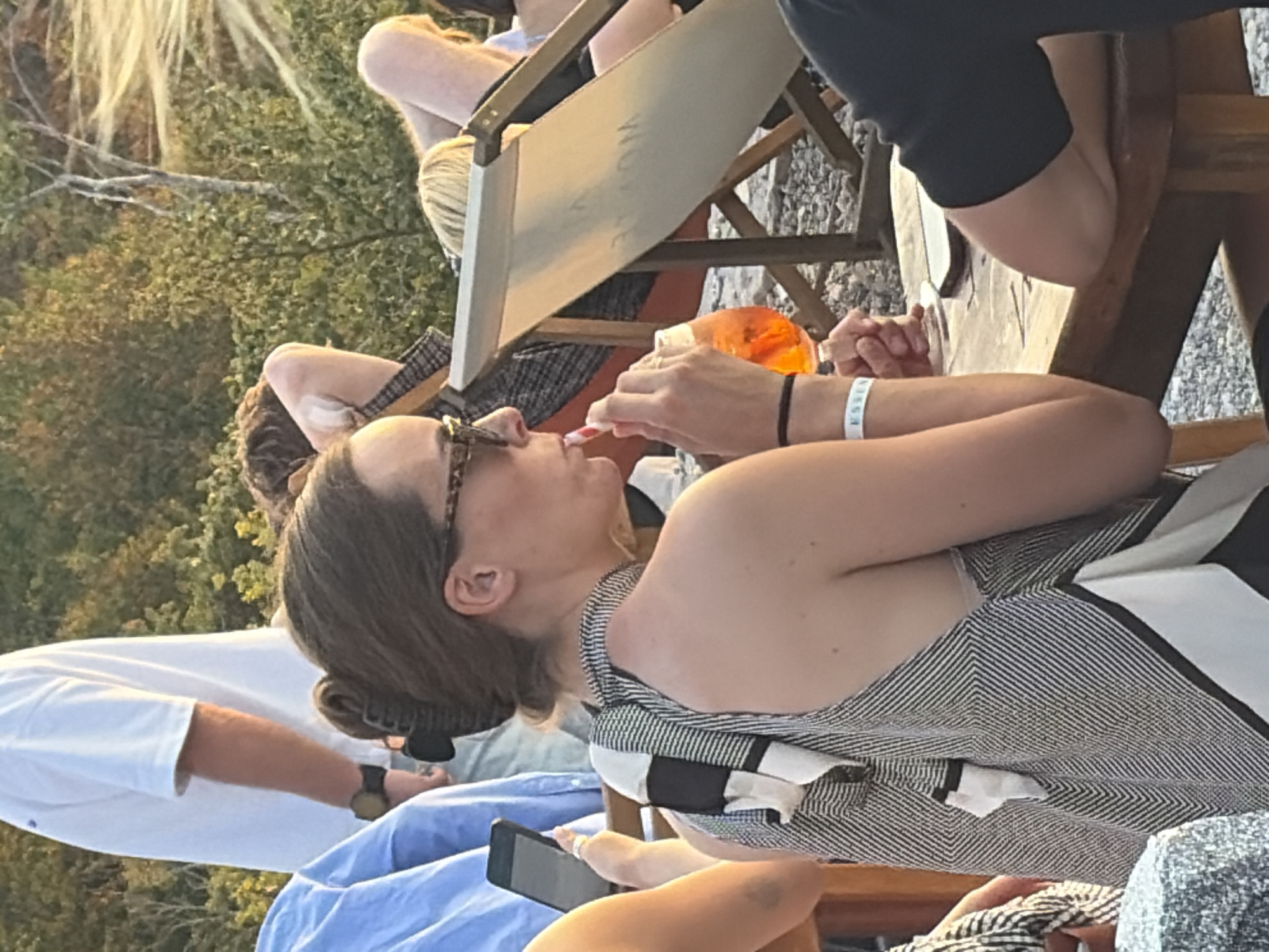 In the city of Kuk, Montenegro, a woman with dark hair pulled into a bun and wearing sunglasses is seated at a wooden table outdoors. She is wearing a black and white patterned halter top and sipping from an orange drink with a striped straw. A white wristband with the word "ESSEN" is visible on her left arm. To her left, another person's arm is visible holding a smartphone. Behind her, several people are relaxing in lounge chairs amidst lush green foliage. The lighting suggests it is late afternoon or early evening.
