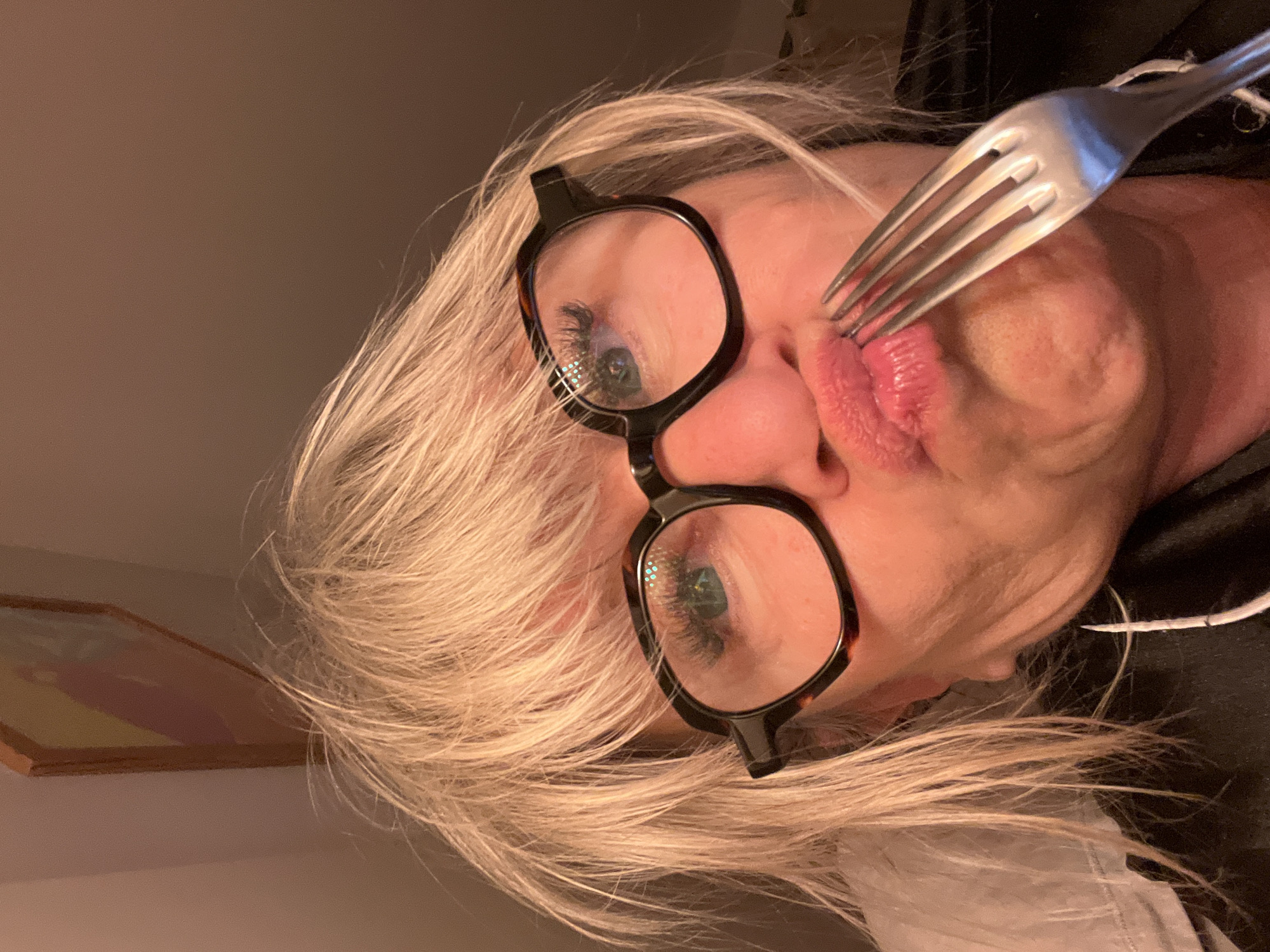 A woman with short, light blonde hair and tortoise-shell framed glasses is holding a silver fork to her pursed lips. The scene is indoors. A piece of artwork hangs on the wall in the background. The location is not discernible.