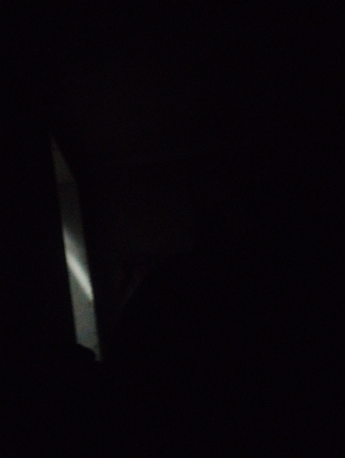 A dimly lit image reveals a corner of what appears to be a building or structure. A vertical, white highlight suggests a sharp edge or corner, possibly illuminated by a source outside the frame. The remaining area of the image is dominated by deep shadows, obscuring detail. A faint reddish hue appears in the center, although it is difficult to discern what it represents.