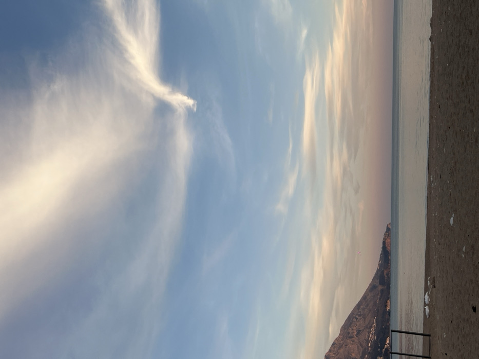 This image depicts a coastal scene in Boreti, Montenegro, observed at either dawn or dusk. The sky occupies approximately two-thirds of the frame, displaying a gradient from light blue at the zenith to pale pink and orange hues near the horizon. Wispy, elongated cirrus clouds are scattered across the sky, with a prominent, bright white, 'swoosh'-shaped cloud formation visible in the upper right quadrant.
The calm, light blue-grey sea spans the middle ground, its surface flat and reflective, meeting a sandy, light brown beach in the foreground. Scattered white debris, consistent with plastic bags or litter, is visible on the sand, particularly in the lower left portion of the beach near the presumed waterline.
To the far left of the foreground, a dark, partial metal structure, resembling a goalpost or frame, is present, with an upright and crossbar visible.
In the background on the left, a large, dark, rocky mountain range slopes down towards the sea. The lower slopes of this mountain are dotted with numerous small, white-walled buildings featuring red roofs. A distinct headland extends from the mountain into the sea, curving slightly to the right.
Above the sea, to the left of the image center and in front of the headland, a small paraglider with a discernible pink and white canopy is visible in flight. No other individuals or explicit human activities are present on the beach or in the water.