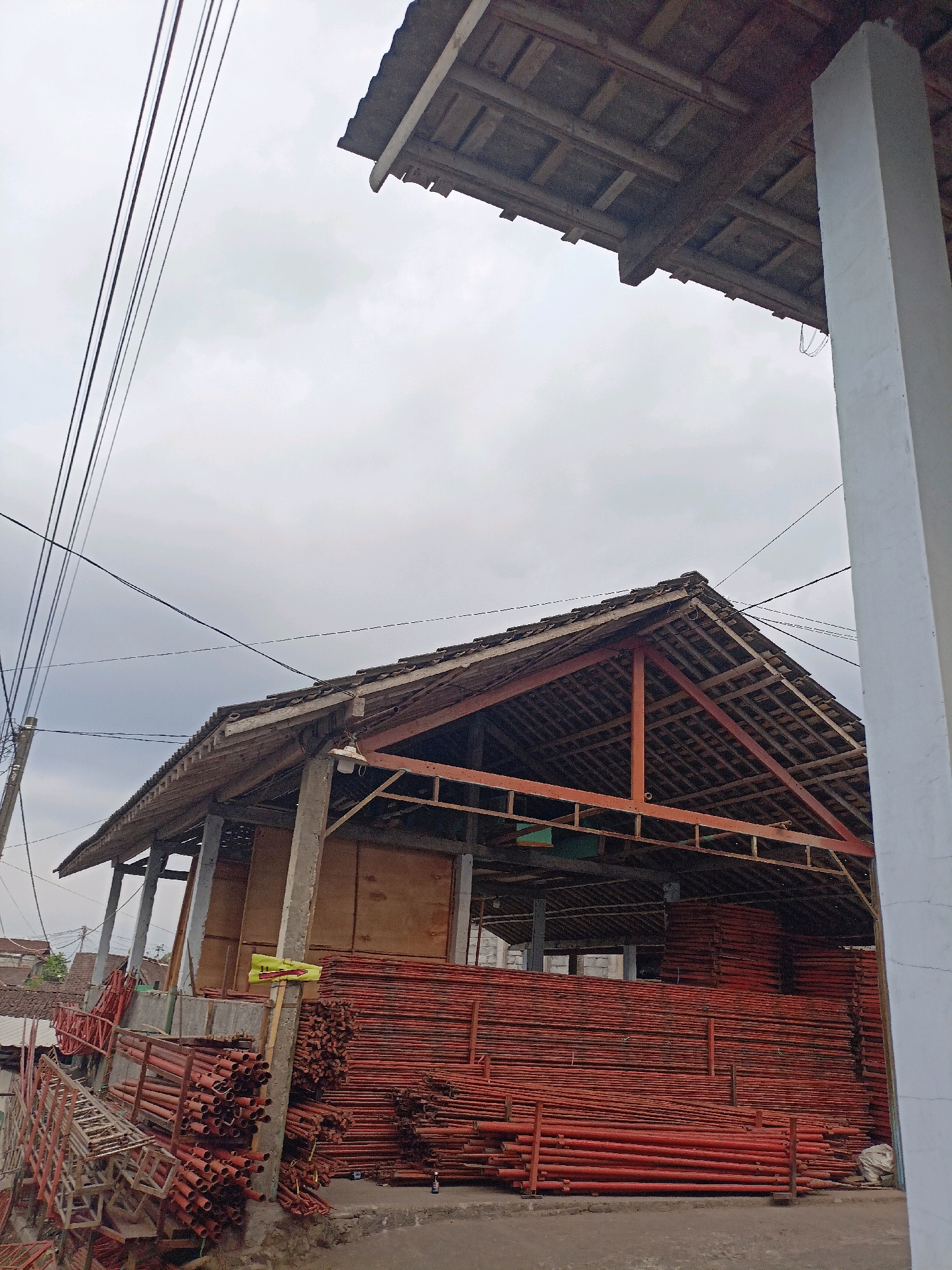 The image depicts a storage area in Bandongan, Indonesia, likely a construction site or a materials depot. In the foreground, numerous red metal poles are stacked in large quantities, some appearing to be scaffolding. To the left, more scaffolding pieces are visible, along with some wooden planks. A partially constructed building with an open roof structure dominates the center of the frame. The roof is covered with tiles and supported by wooden rafters and some metal beams. The walls of the building are constructed from what appears to be wooden planks. Utility wires are strung across the sky above the structure. The ground is paved with concrete or asphalt. The sky is overcast, suggesting a cloudy day. No people are visible in the image.