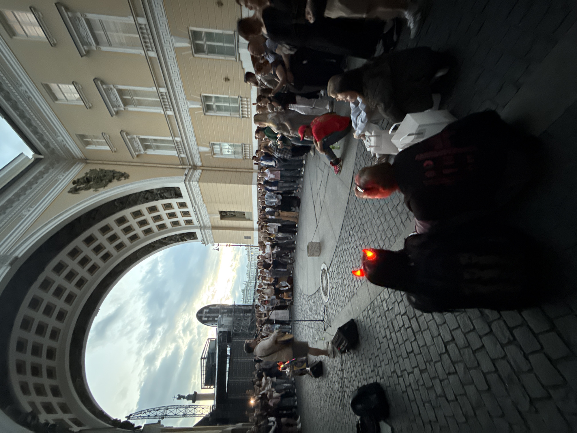 A man wearing a light-colored shirt and shorts is playing an acoustic guitar and singing into a microphone in a cobblestone plaza. He is standing near a portable speaker and several other pieces of audio equipment. A crowd of people of various ages are gathered around him, some standing and some sitting on the ground. In the background, there is a large, ornate archway leading to a pale yellow building with many windows. The sky above is cloudy and appears to be at dusk. Two people in the foreground are wearing what appear to be devil horns with red lights.