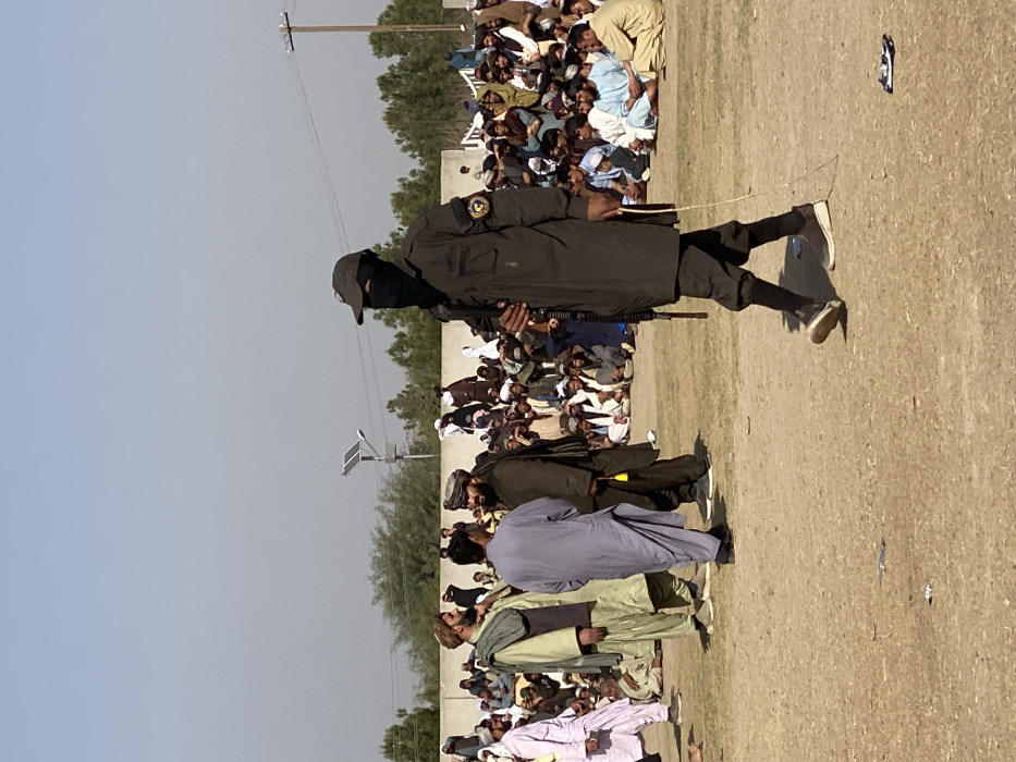 The image displays an outdoor scene in Kandahar, Afghanistan, under a clear daylight sky. The foreground consists of dry, light brown dirt ground, with sparse areas of dry grass and minimal debris, including a crumpled piece of material near the bottom right.
In the midground, a male figure walks from right to left, positioned prominently on the right side of the frame. He wears a dark, long-sleeved uniform, a dark baseball cap, and a black face covering that obscures his facial features. He holds a dark, long-barreled rifle vertically, resting against his right side, and a circular emblem is visible on his left sleeve. A thin, light-colored rope or cord dangles from his left hand. He wears dark athletic-style shoes with white soles.
To the left of this figure, three men stand engaged in conversation. The leftmost wears a light green tunic (shalwar kameez) with a dark vest. The central man wears a light blue tunic. The rightmost wears a dark brown tunic and a dark turban, featuring a yellow tassel. All three men have beards.
Behind these individuals, a large crowd of men is seated directly on the ground, extending into the mid-to-background. They are dressed in various traditional Afghan garments, including multiple colors of shalwar kameez and turbans, appearing to observe an event. In the background, a low, light-colored concrete or stone wall is visible, backed by a line of green trees. Two utility poles, one fitted with a solar panel and a streetlight, rise against the clear light blue sky.