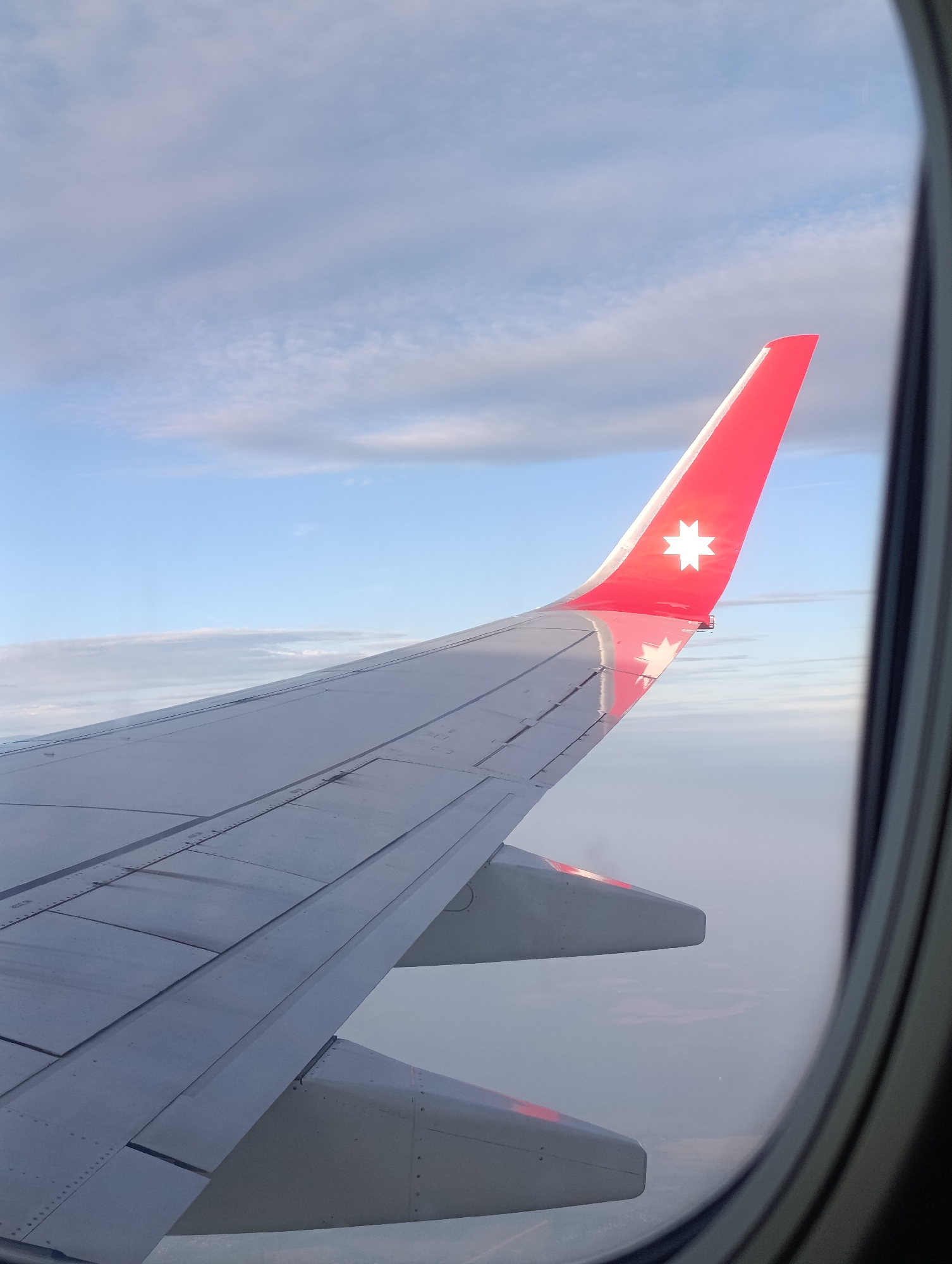 The image shows the wing of an airplane seen from inside the cabin, looking out a window. The wing is predominantly grey and white with a red tip featuring a white star emblem. The sky is visible through the window, with patches of blue and wispy white clouds, suggesting daytime. The scene appears to be during an airplane flight, with the vast expanse of sky and clouds indicating a high altitude. The perspective from the window frames the wing and the sky, creating a sense of travel and perspective. There are no people visible in the frame, and the focus is on the airplane's wing and the surrounding sky. The lighting suggests it could be early morning or late afternoon, given the soft light on the clouds and wing. No visible text is present in the image.